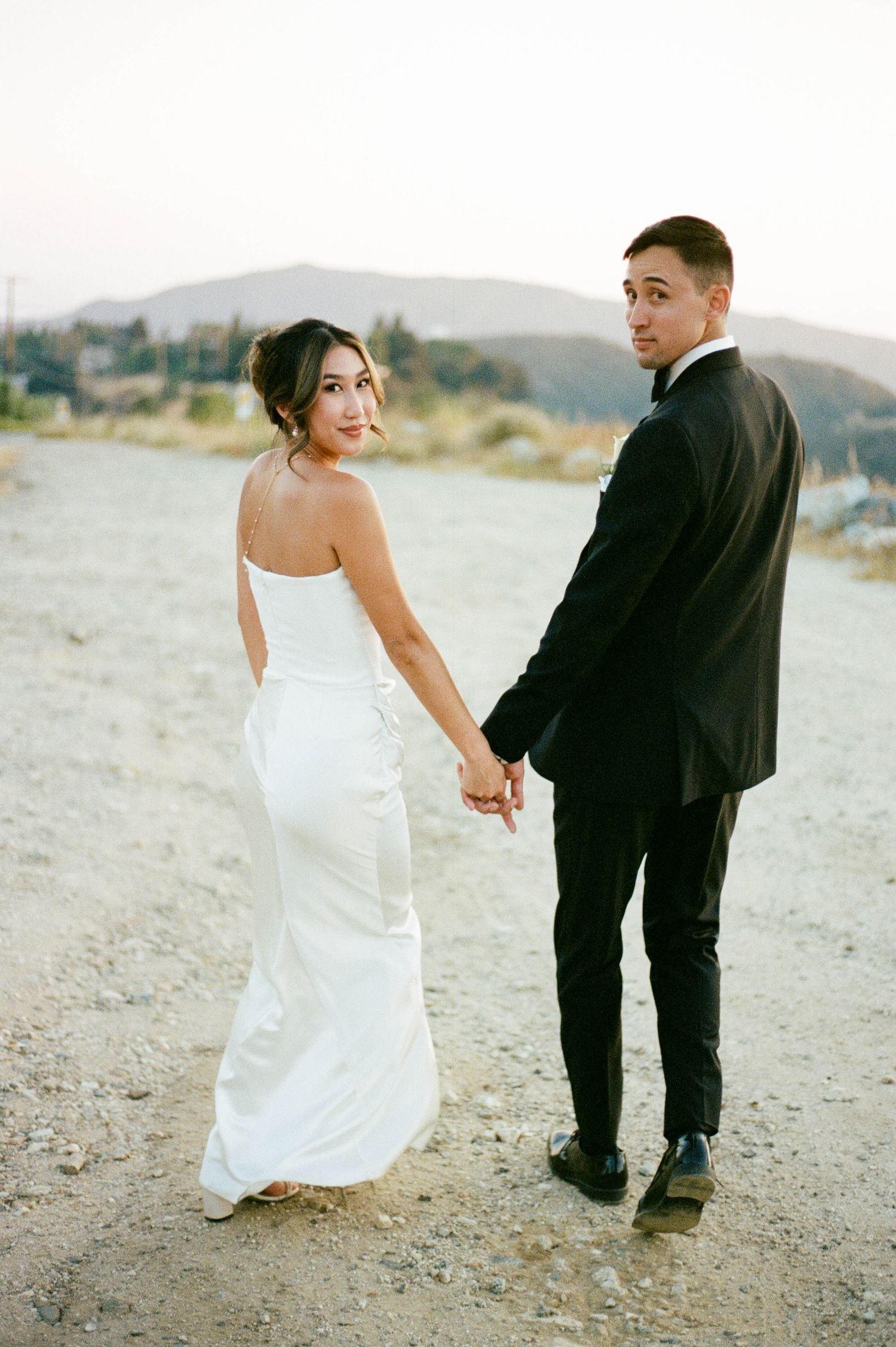A couple takes romantic wedding photos walking together on a sandy beach with mountains in the background.