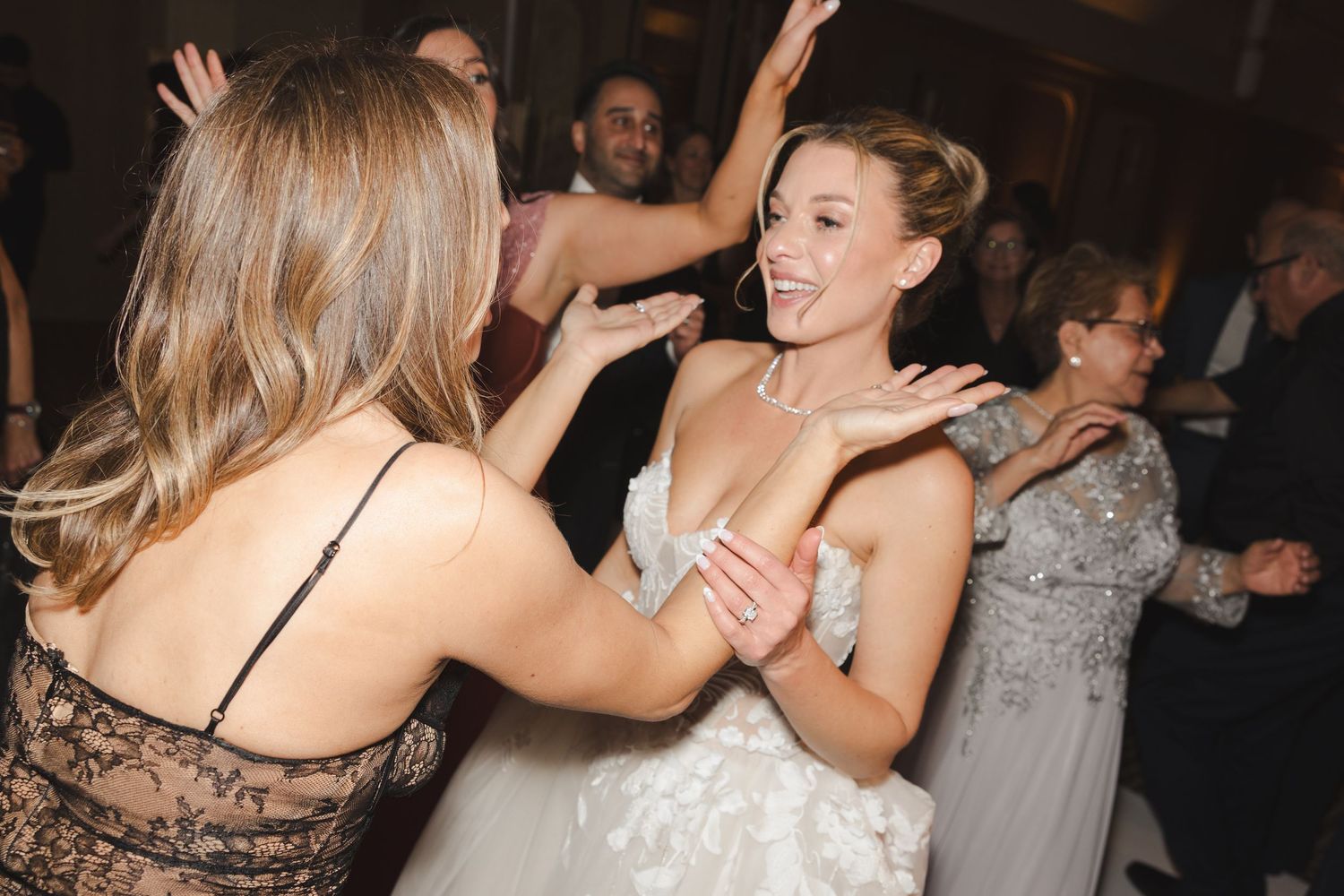 Two people dance together joyfully at a wedding reception, with one wearing a white wedding dress and another in black lace.