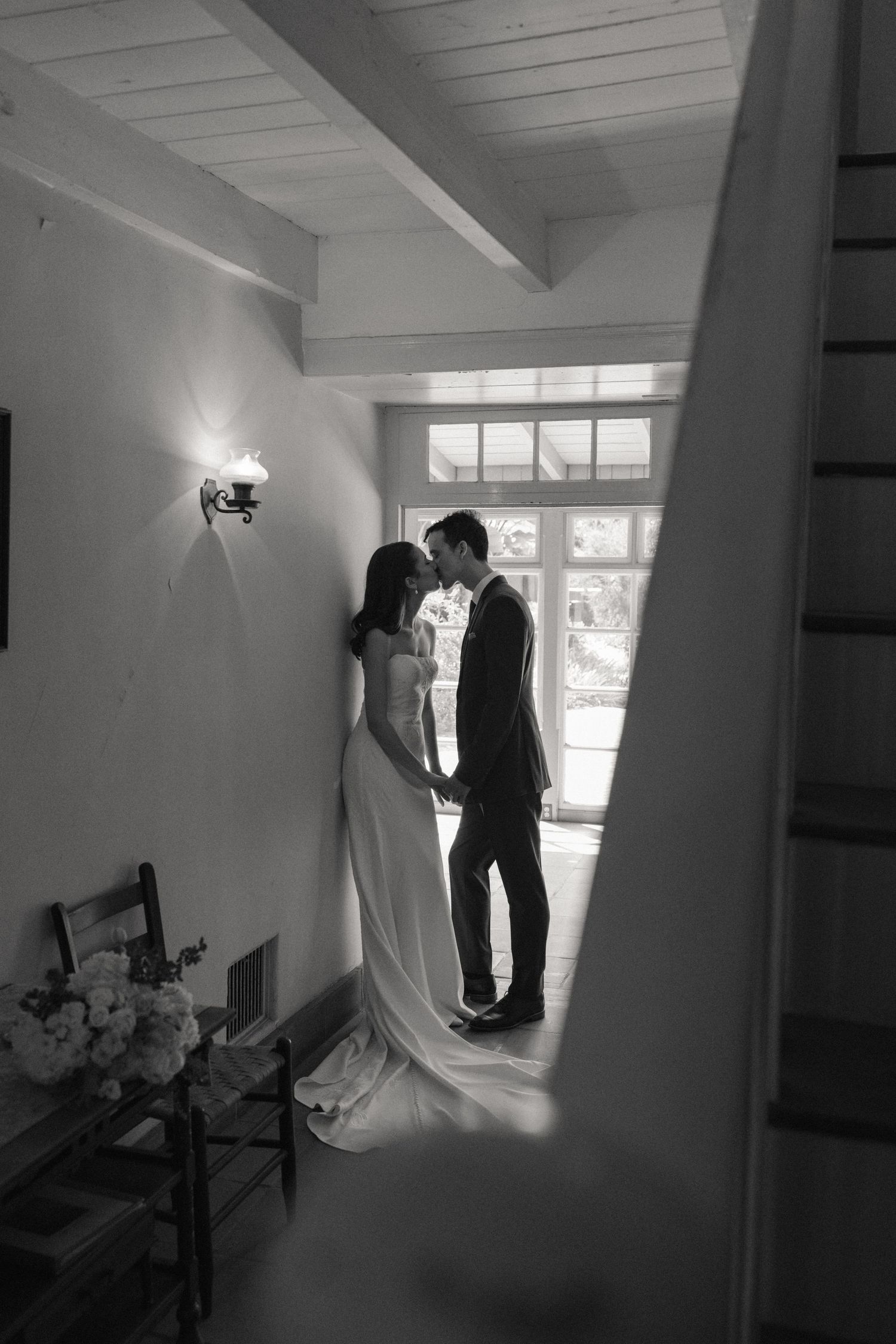 Black and white photo of a bride and groom sharing an intimate moment indoors, framed by a staircase.