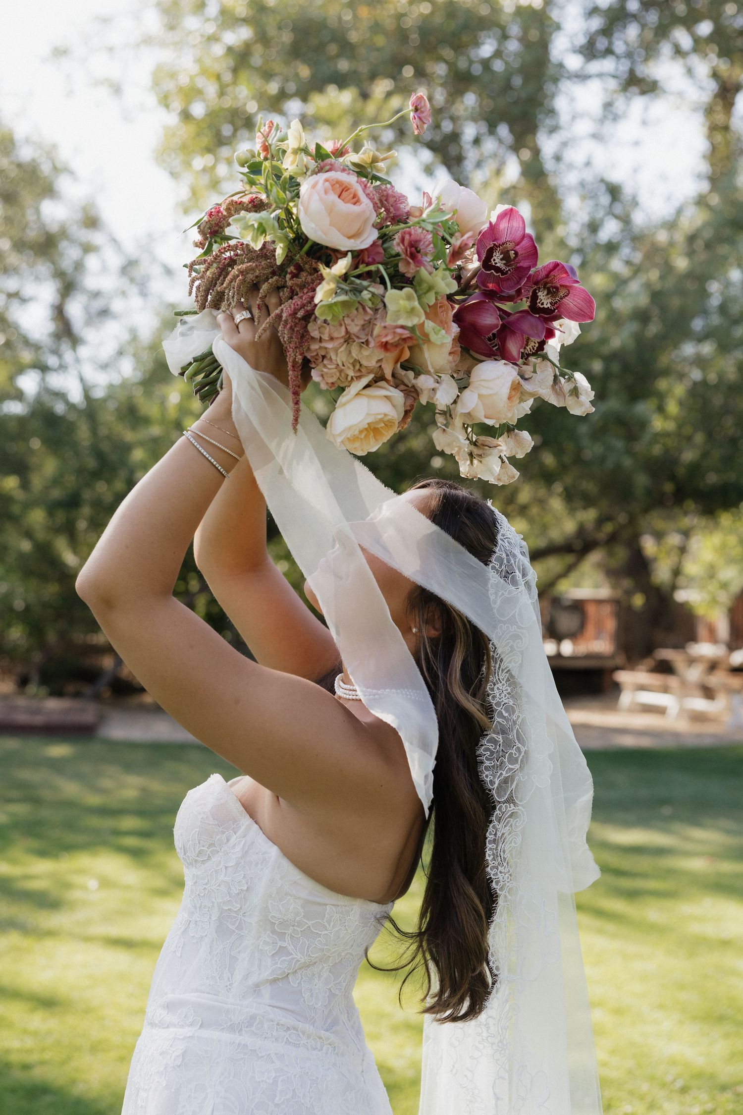 Bride in white lace gown holds colorful floral bouquet overhead with veil flowing outdoors.