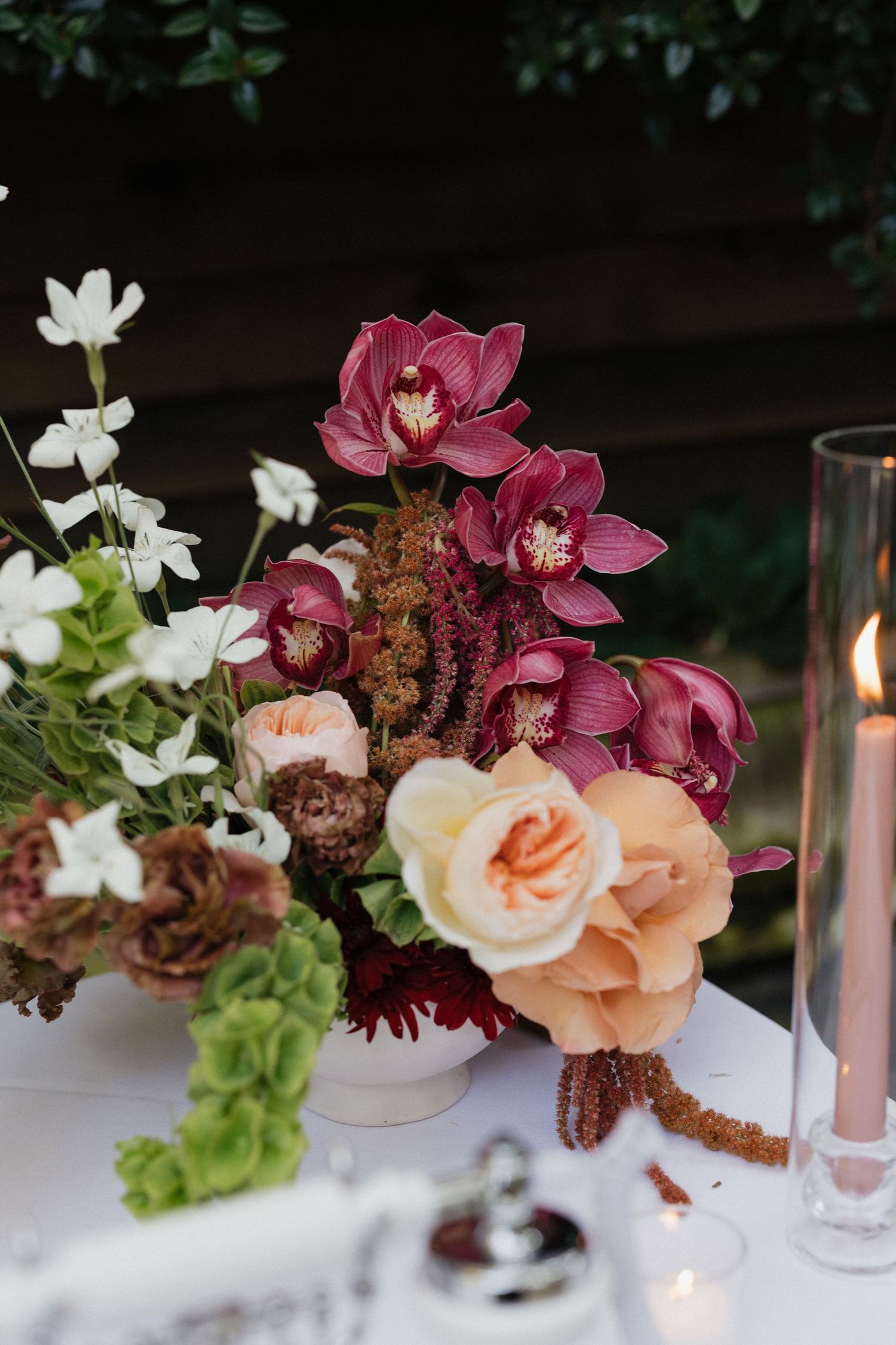 Elegant wedding centerpiece with burgundy orchids, peach roses, and white blooms in white bowl beside candle.