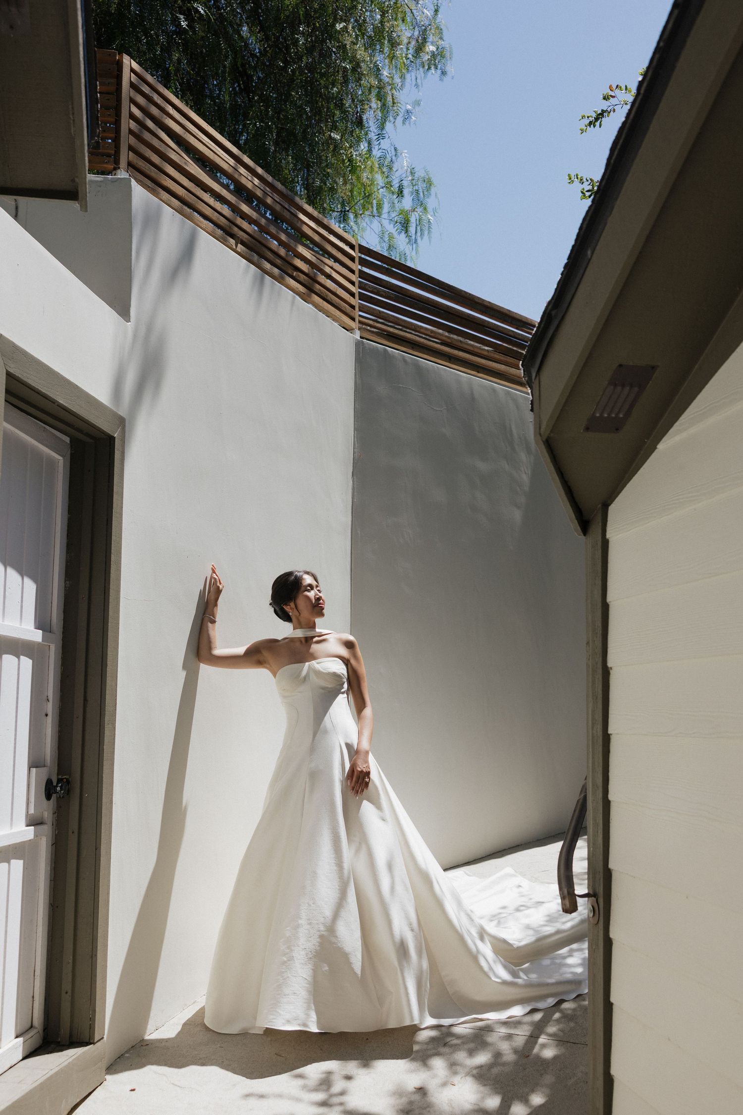Bride in elegant white ball gown with long train leaning against white wall in sunlit outdoor space.