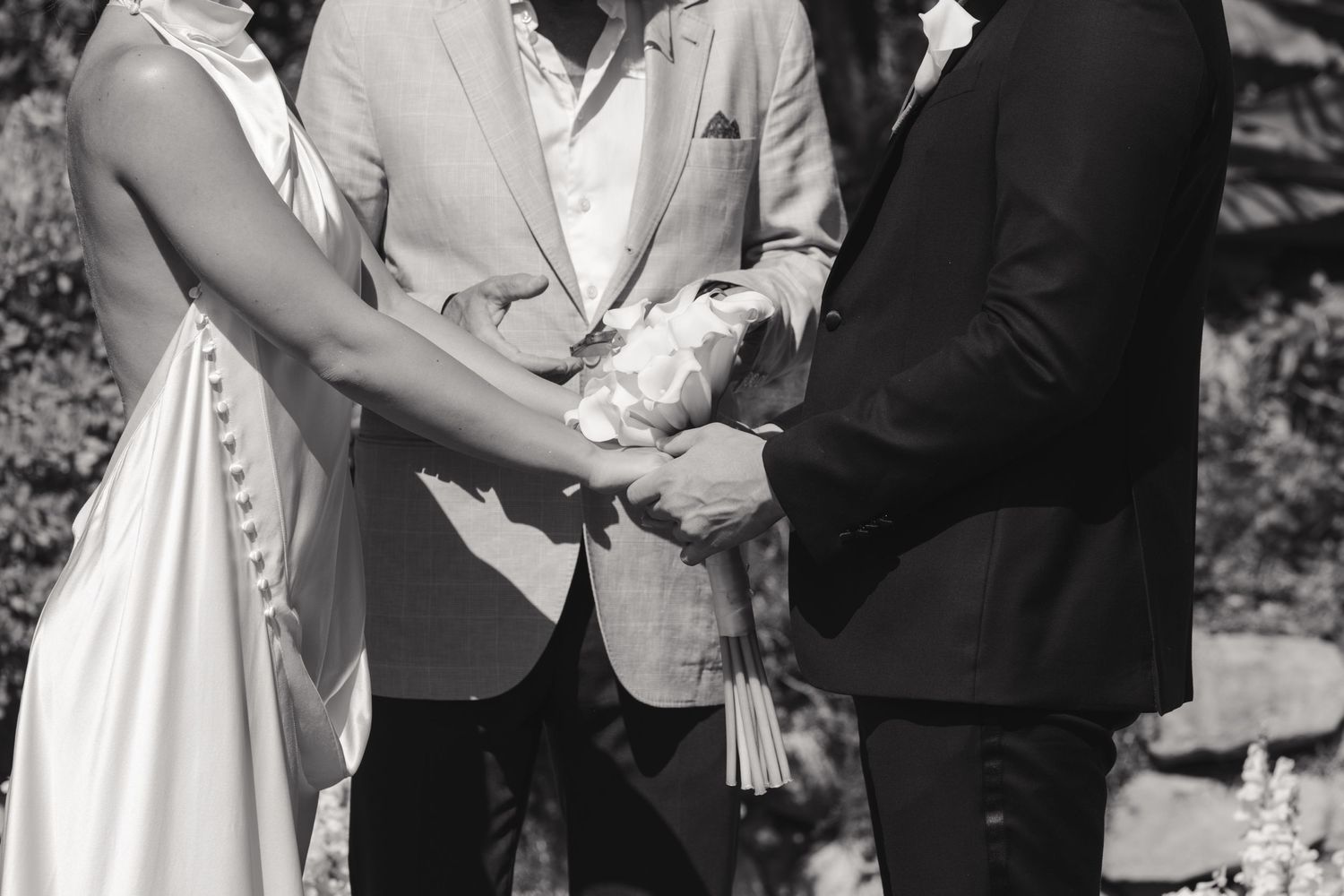 A black and white photograph capturing hands joined together during an outdoor wedding ceremony.