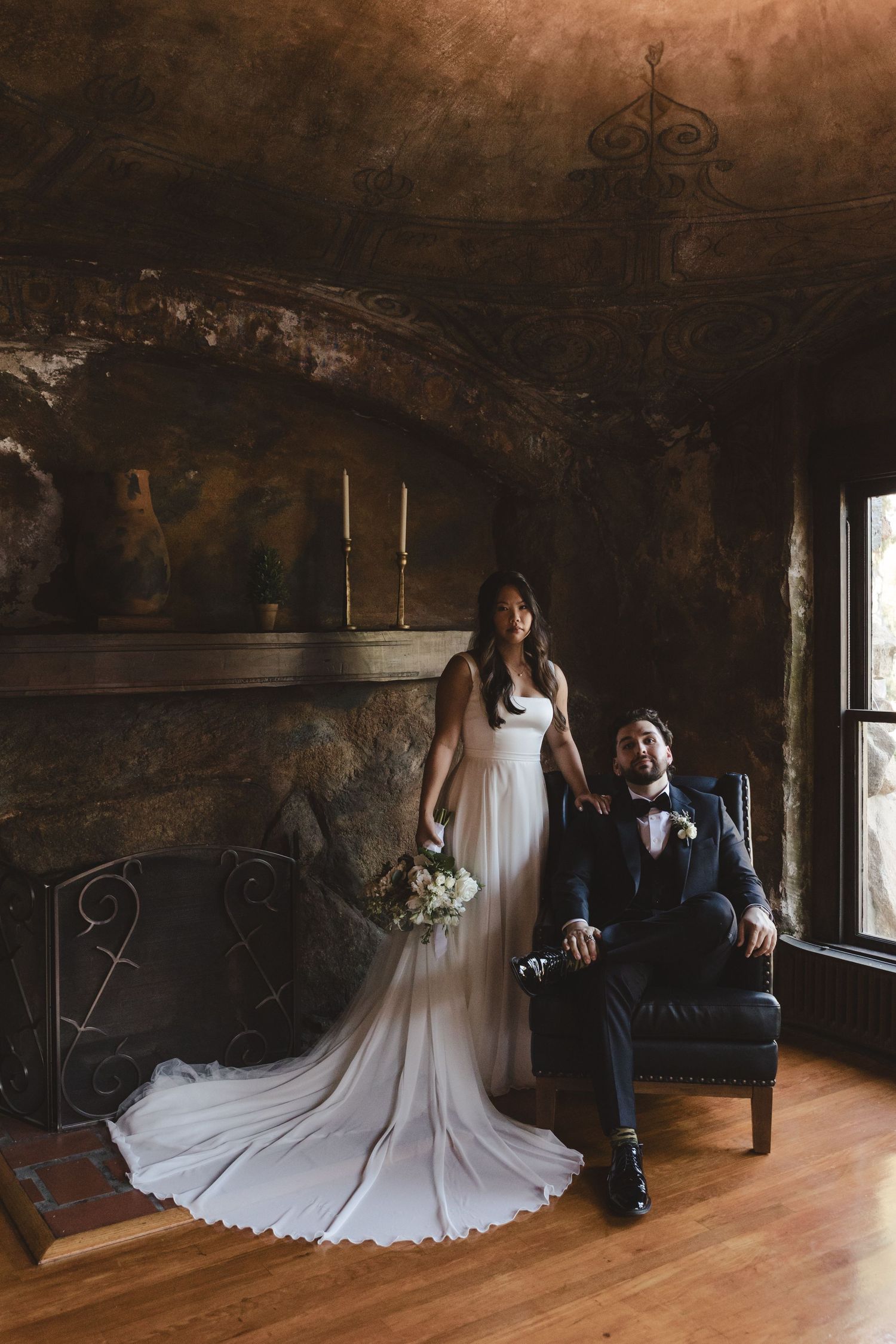 Elegant wedding couple poses in rustic venue with stone walls, dark moody lighting, and hardwood floors.