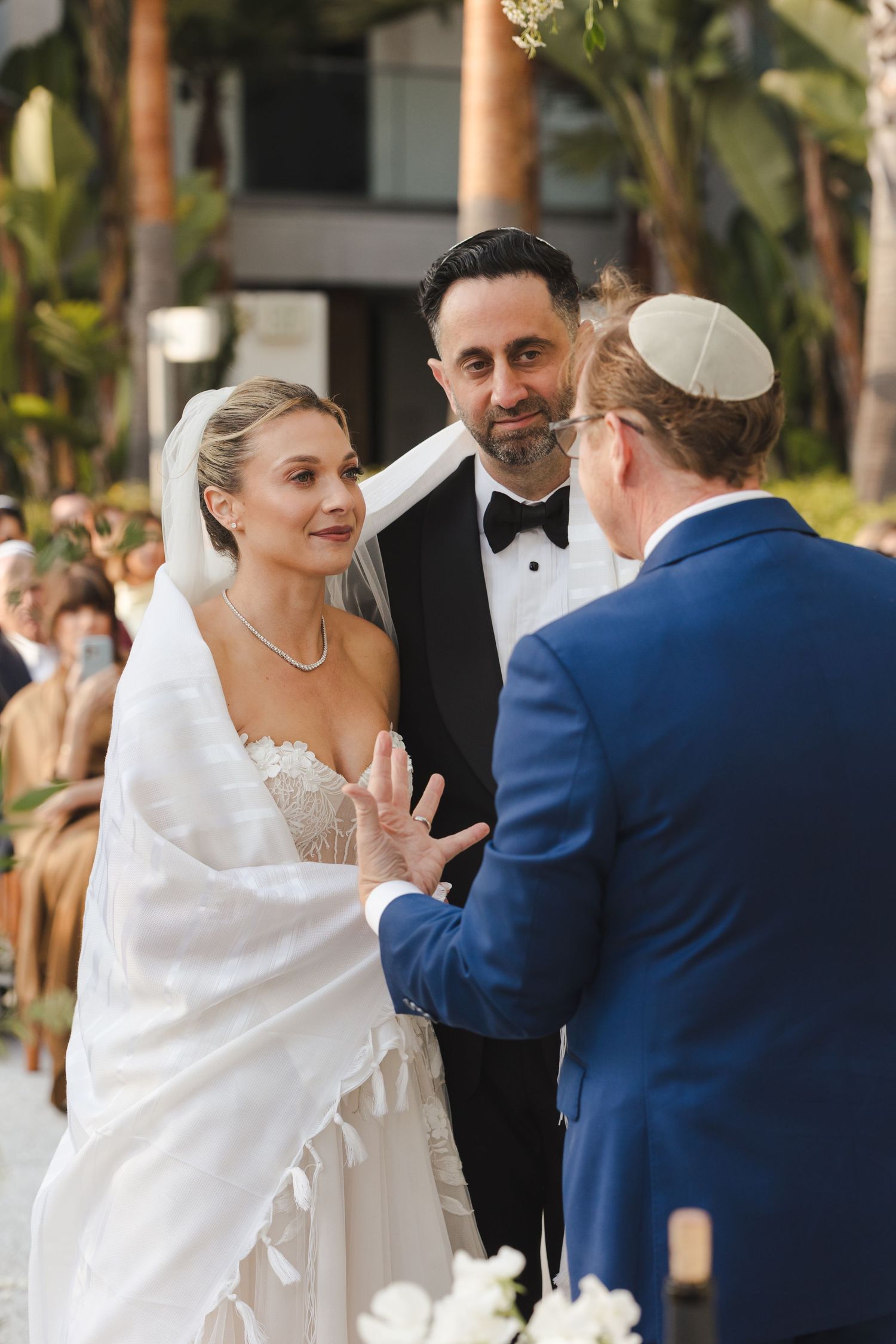 Wedding ceremony takes place in a tropical garden setting with palm trees in the background.