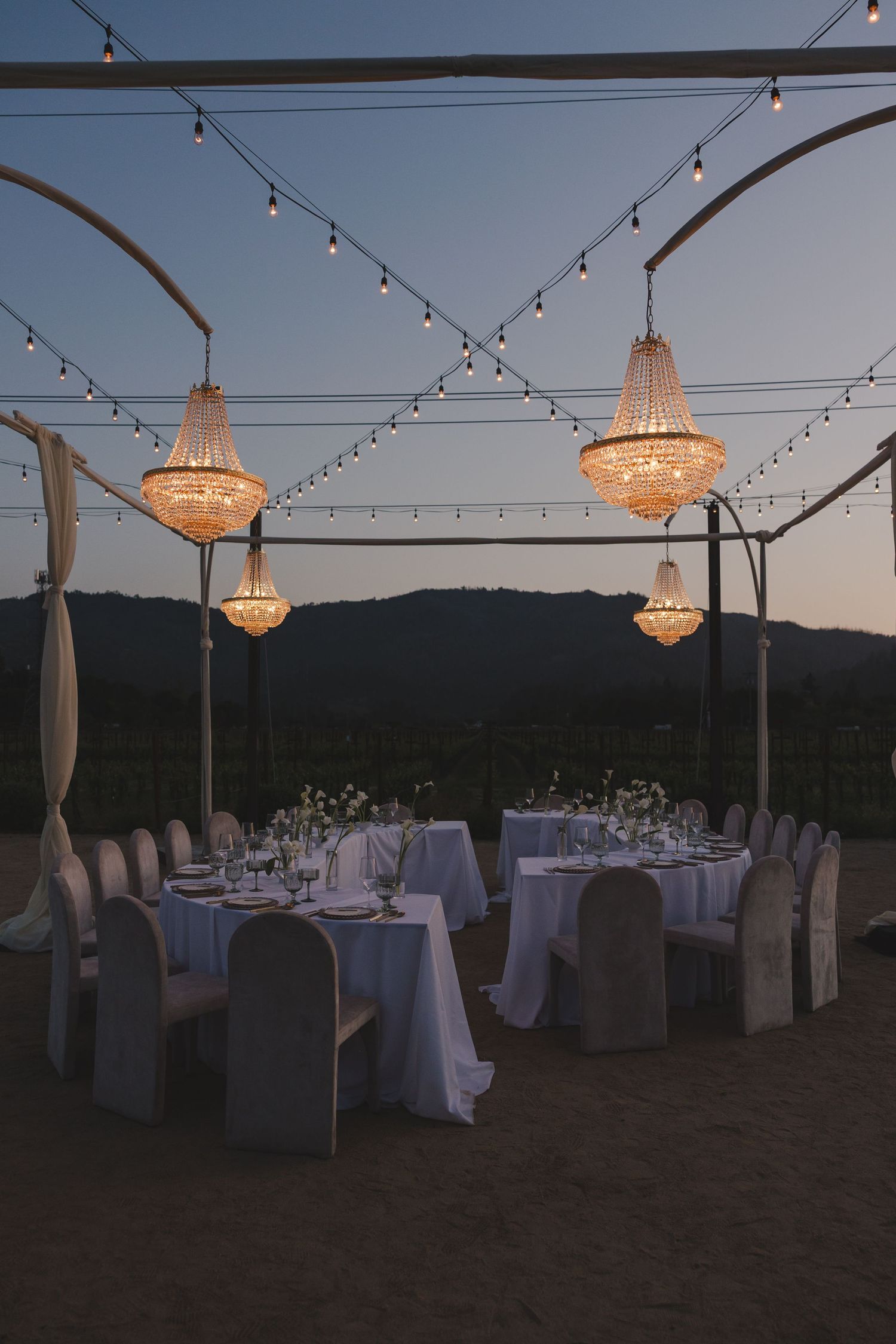 Elegant outdoor wedding reception setup with crystal chandeliers and string lights illuminating tables at dusk.