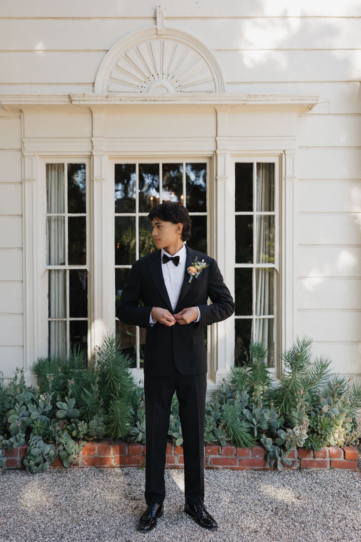 Formal wedding attire detail shot against white colonial style house with brick foundation and greenery.