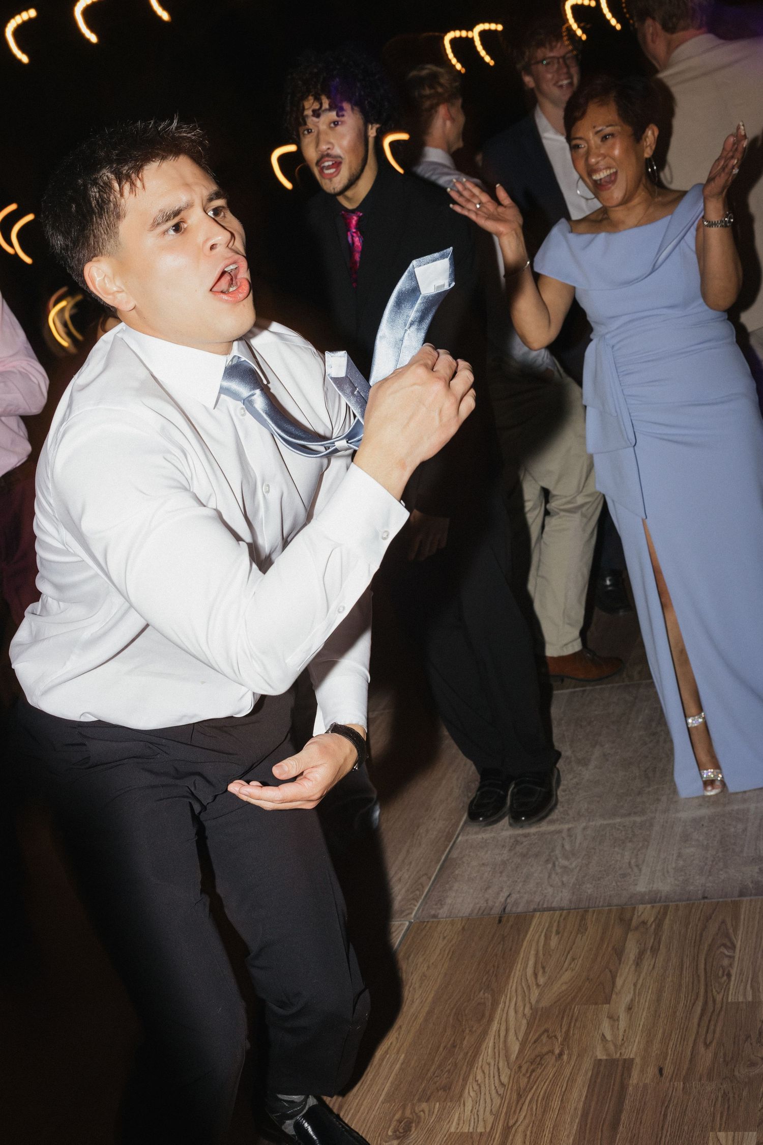 Young guests dancing and singing energetically at a wedding reception on a wooden dance floor.