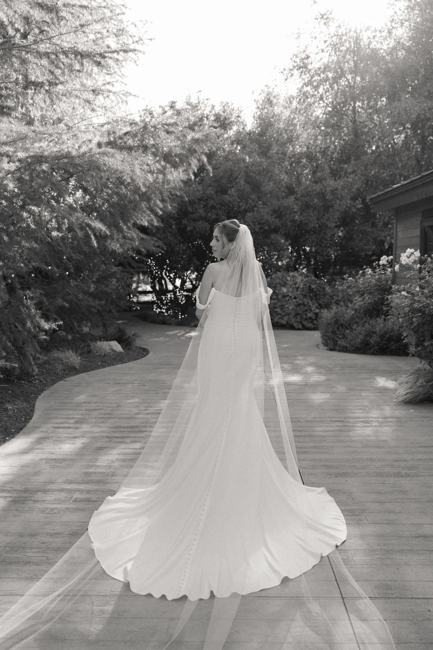 A flowing white wedding dress and veil captured from behind on a garden pathway.