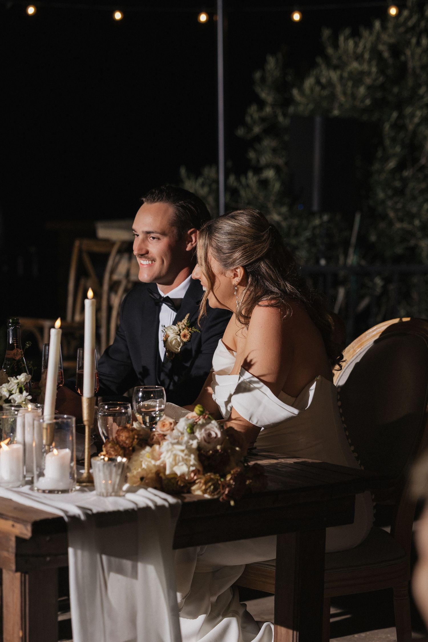 A couple sits at an elegant candlelit reception table with white linens and floral centerpieces at an outdoor evening event.