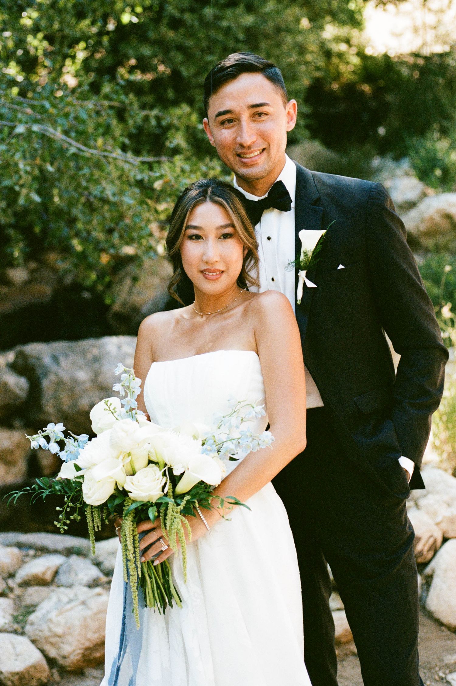 Outdoor wedding portrait with elegant couple holding white floral bouquet against rocky natural backdrop.