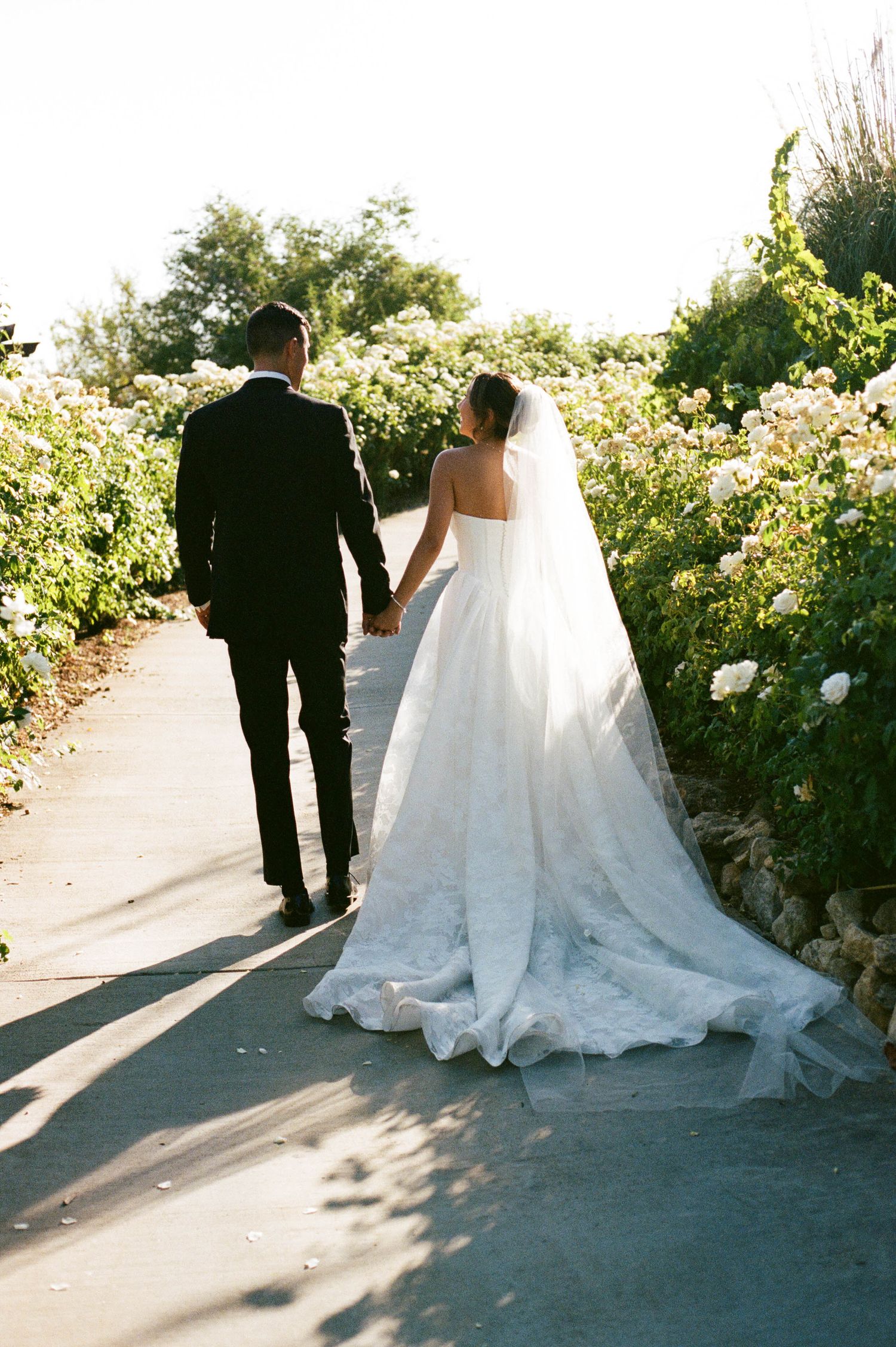 Wedding couple walking along garden path with bride's elegant white dress trailing behind.