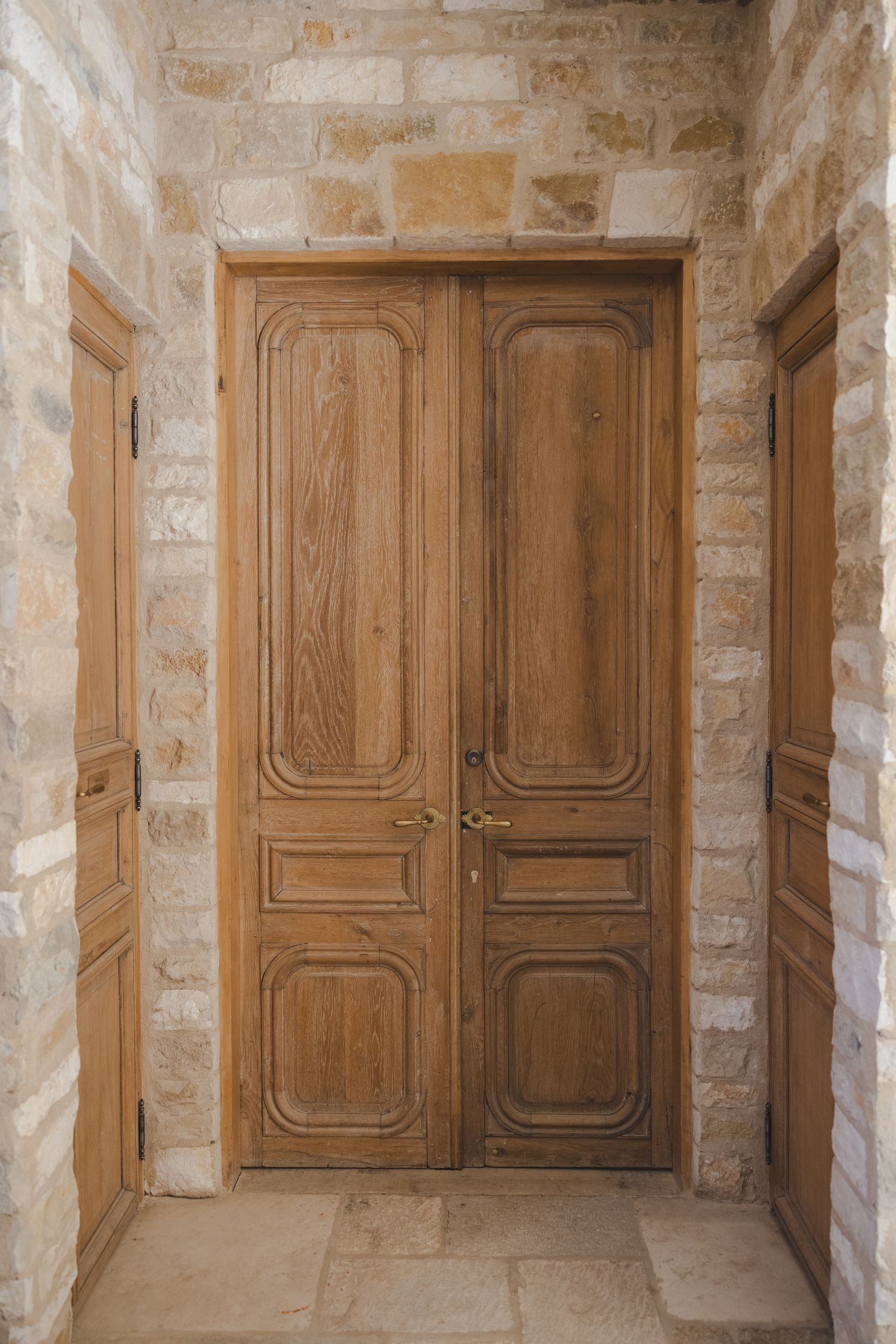 A vintage wooden double door with carved panels set in a stone brick hallway with natural lighting.