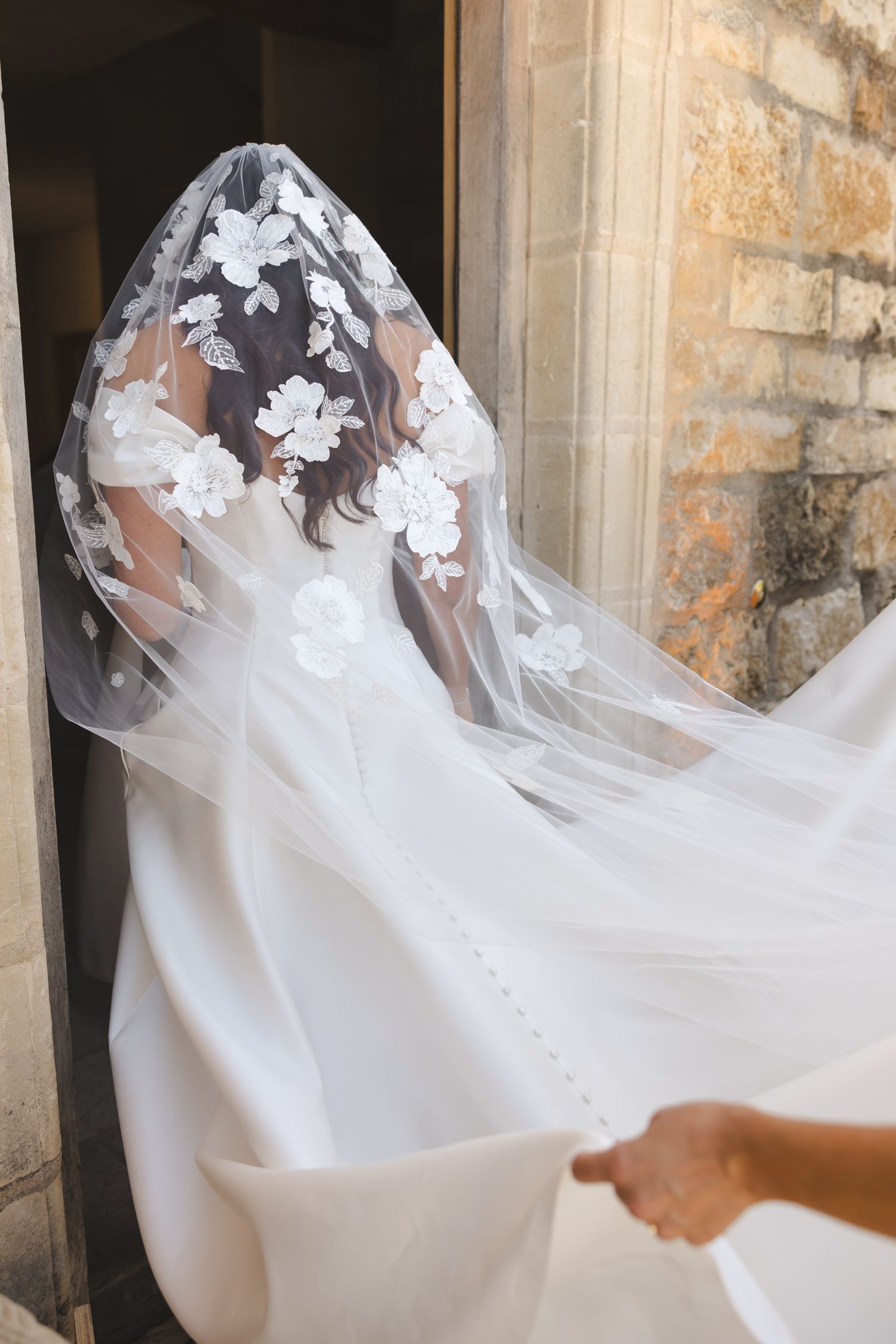Elegant white wedding dress with sheer floral lace veil captured in a stone doorway.