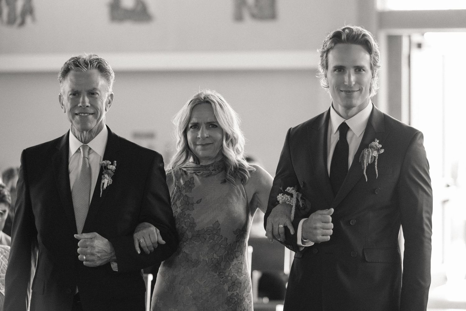 Black and white photo of a bride walking down the aisle escorted by two men in suits at a wedding ceremony.