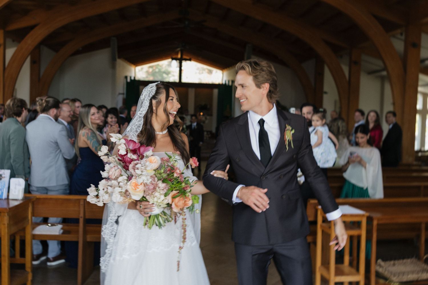 Bride and groom walk down the aisle smiling, bride holding a lush floral bouquet in a rustic wood-beamed chapel.