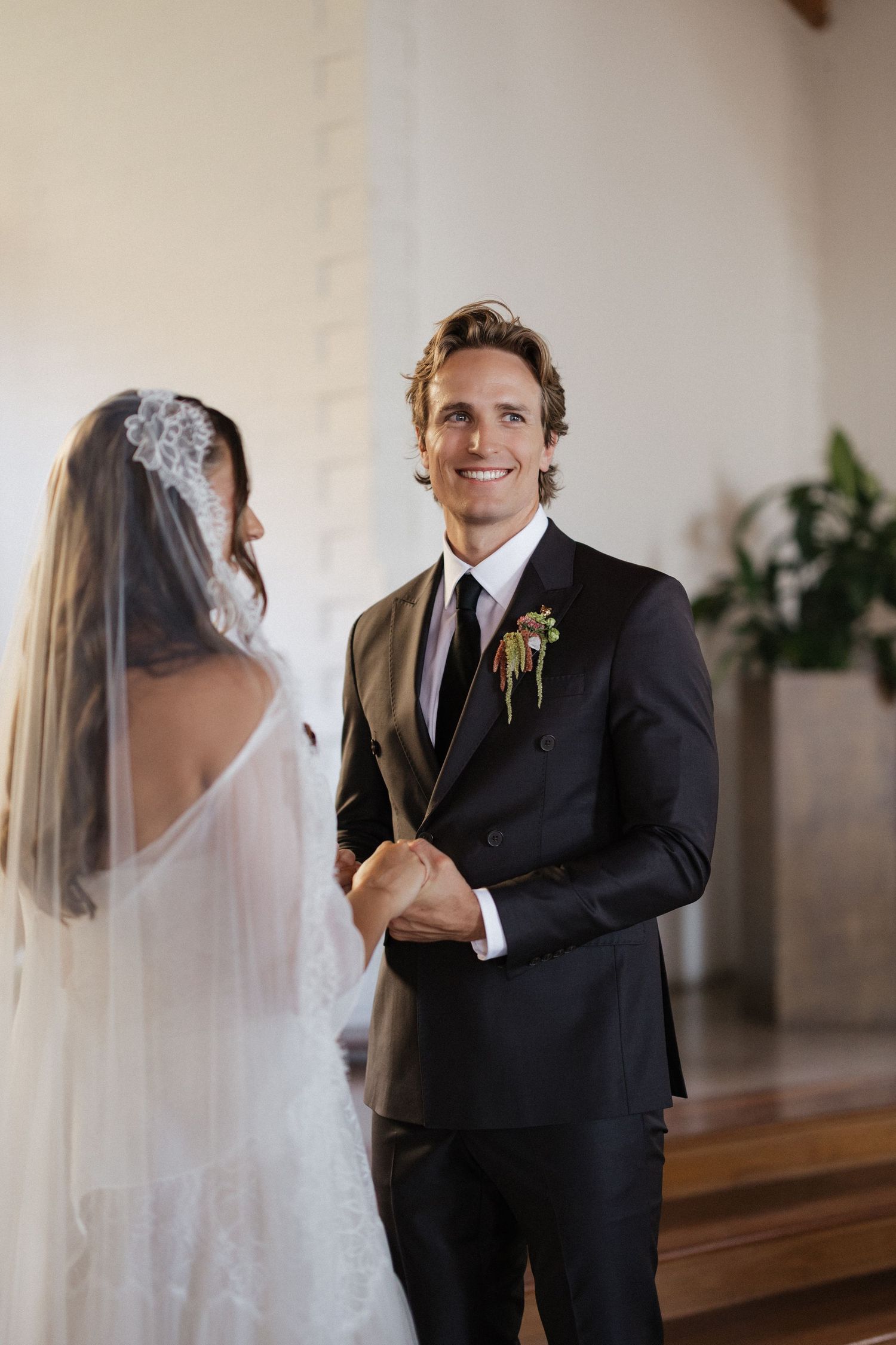 Smiling groom in black suit holding bride's hands at wedding ceremony altar.