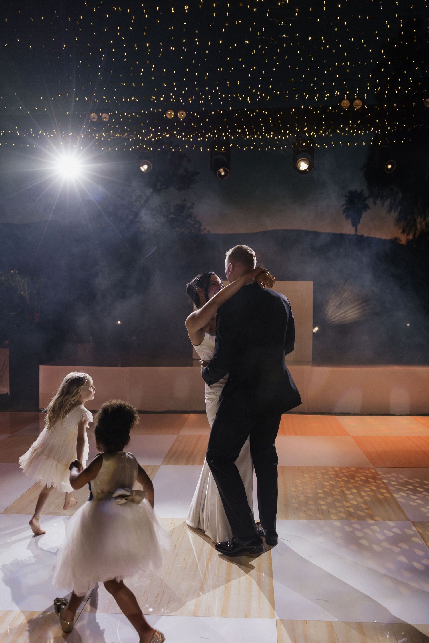 Bride and groom share first dance under twinkling lights with flower girls on elegant wedding dance floor.