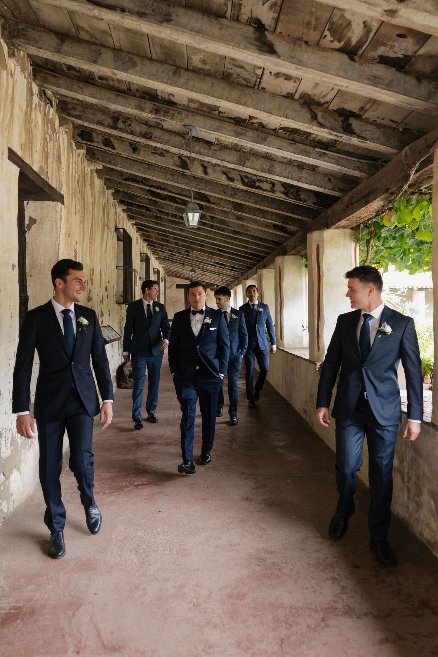 Groomsmen in navy suits walk together down a rustic covered corridor at a wedding venue.