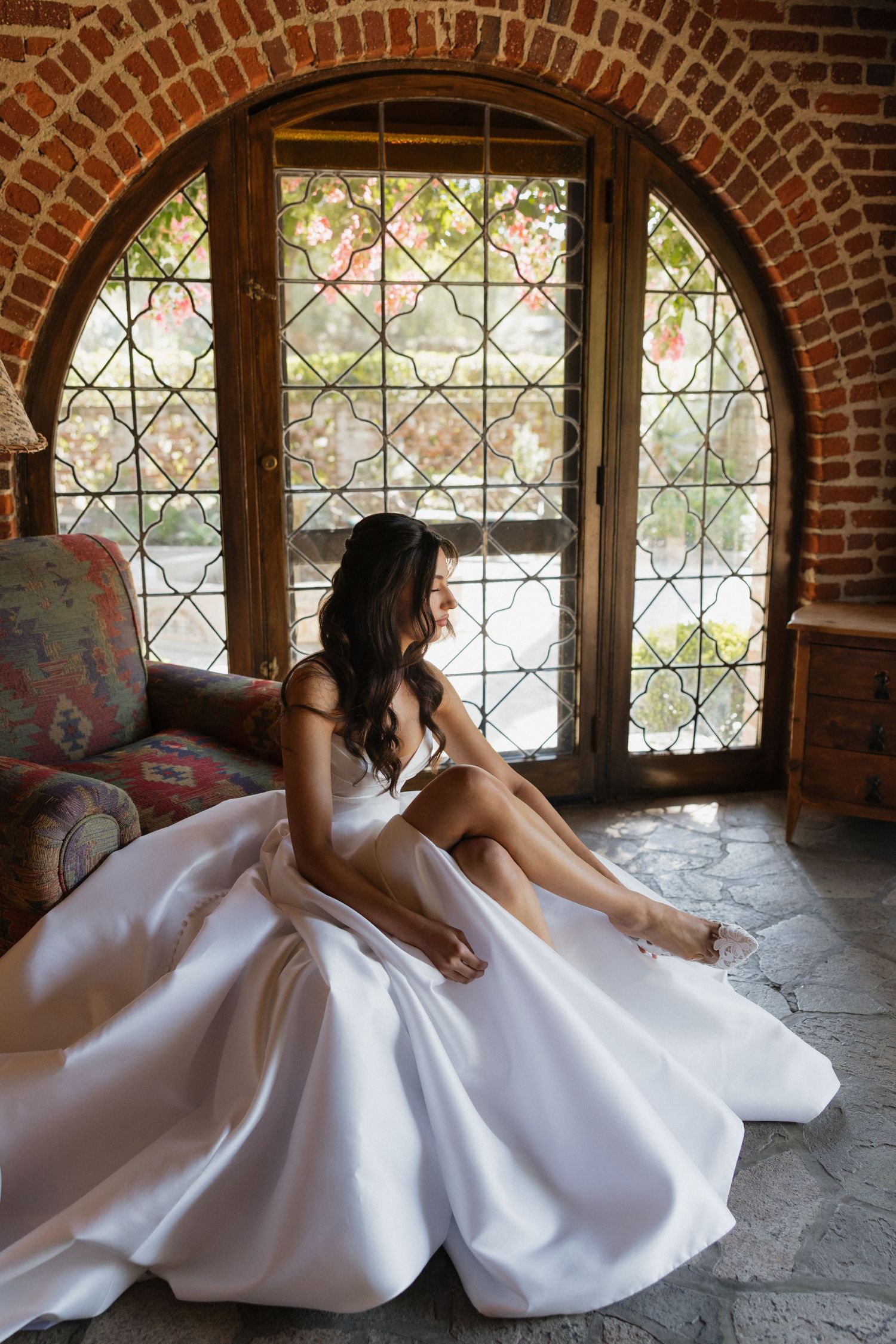 A bride in a white gown poses gracefully near arched brick windows with decorative lattice details.