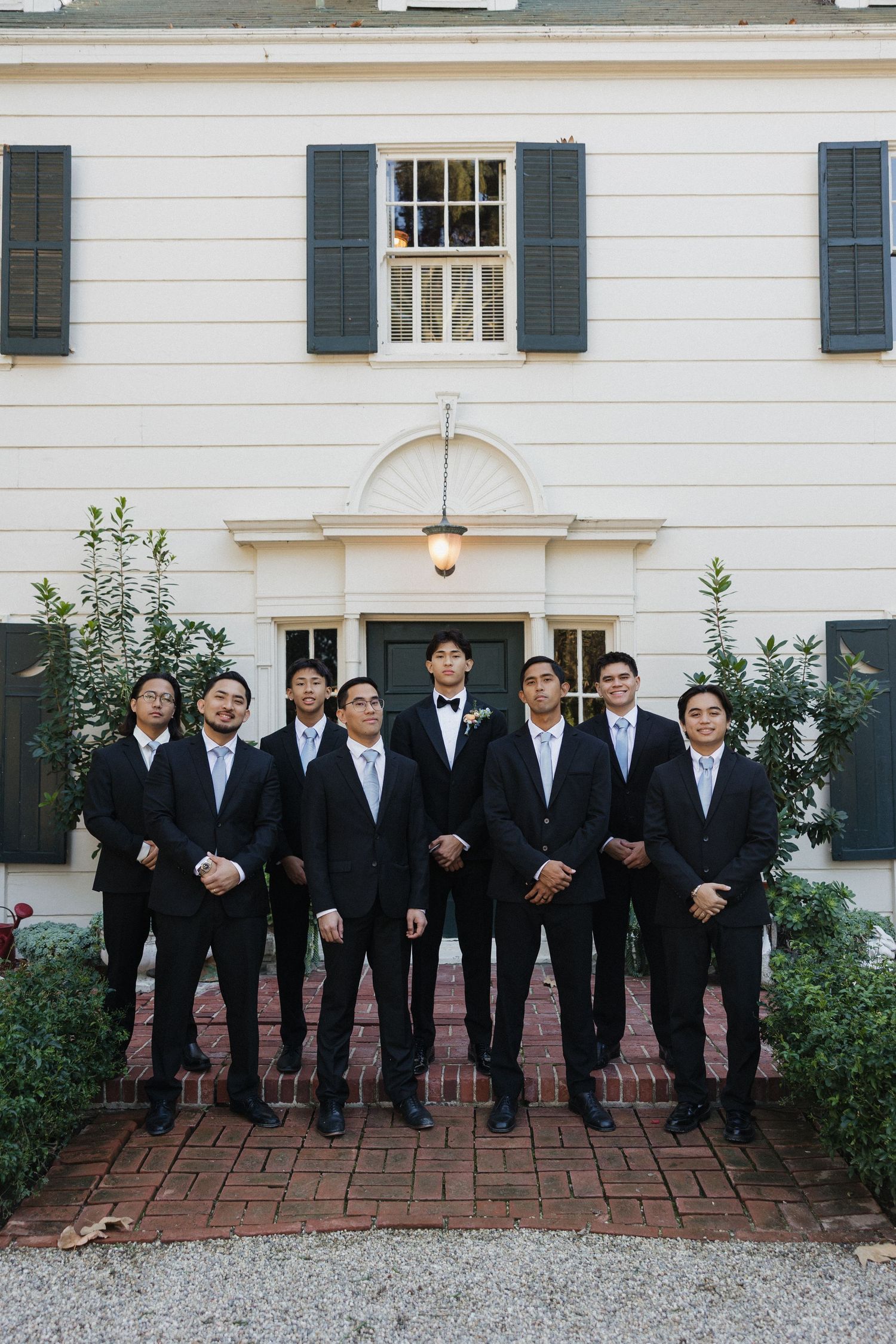 Groomsmen in black tuxedos line up along brick pathway in front of classic white colonial building.