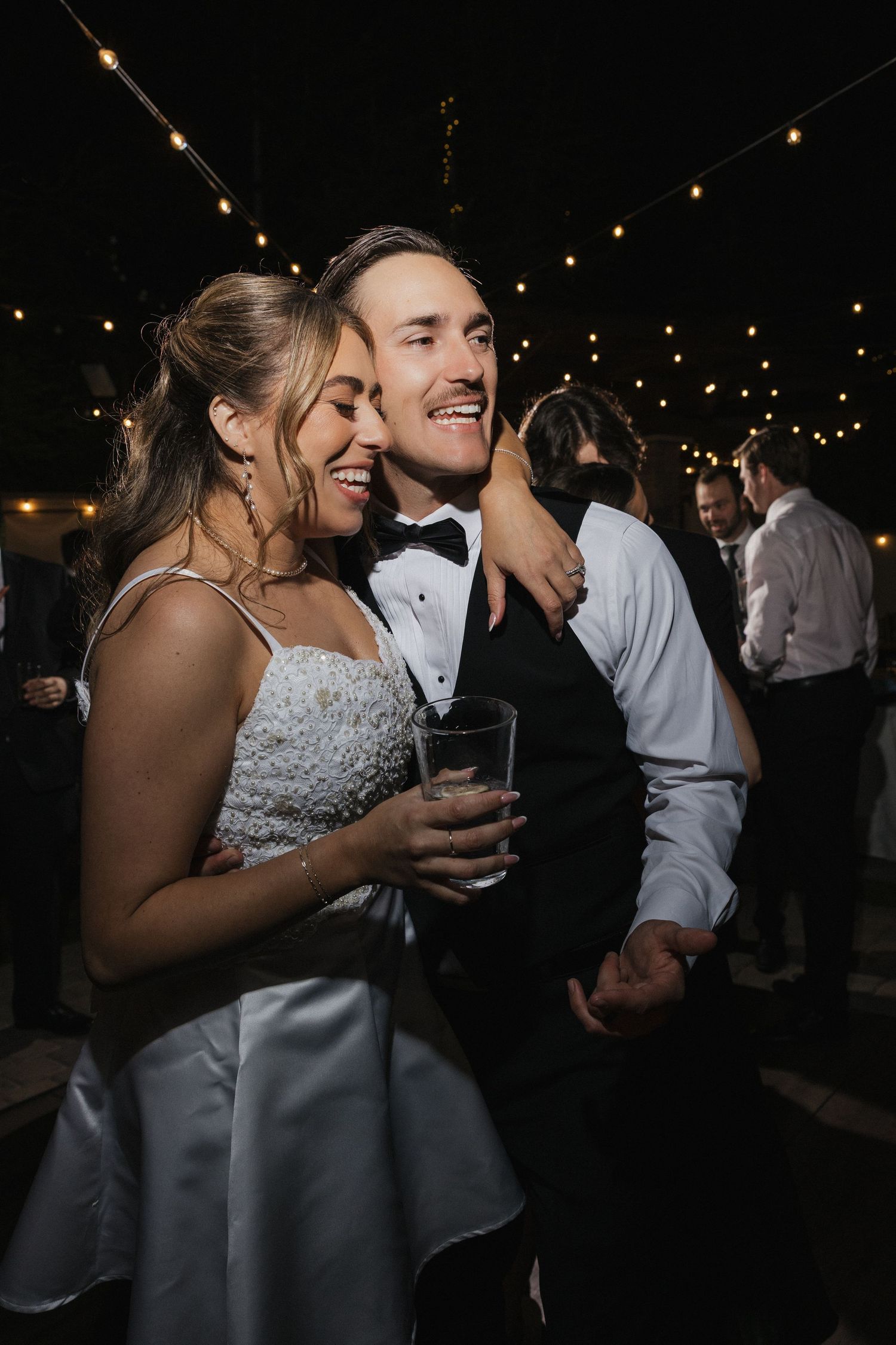 A couple shares a joyful moment at their wedding reception under string lights, with the bride wearing a white gown.