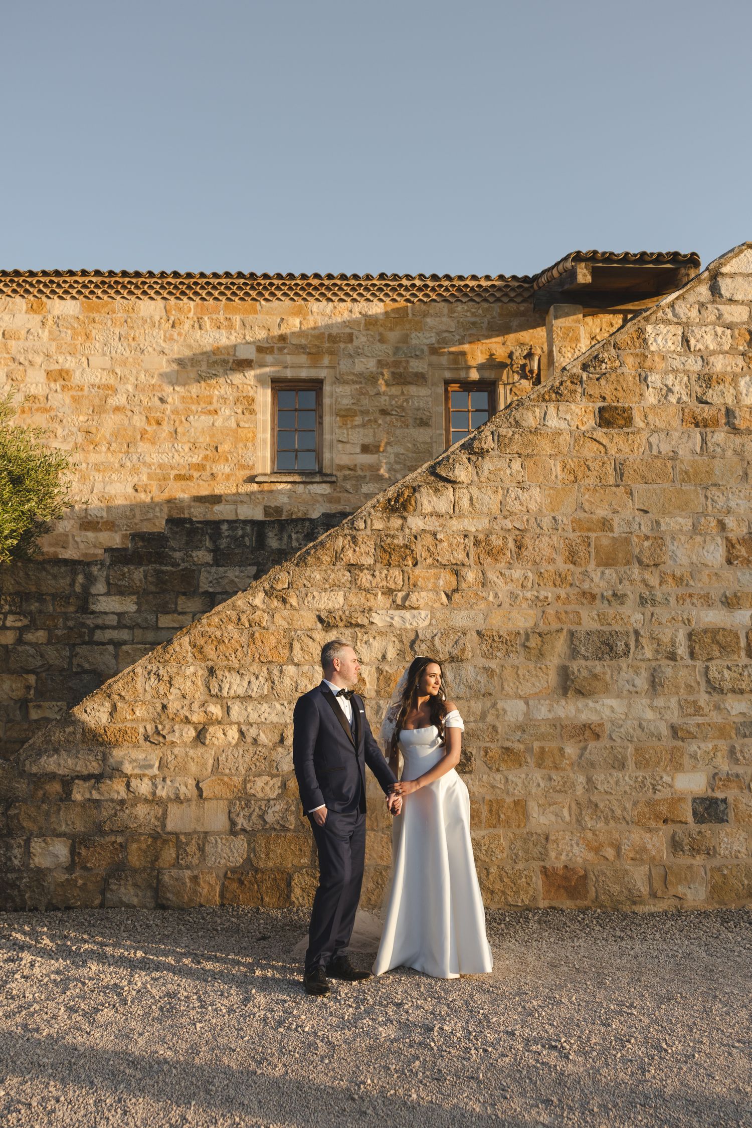 A couple stands against a rustic stone wall at sunset casting long shadows across the textured facade.