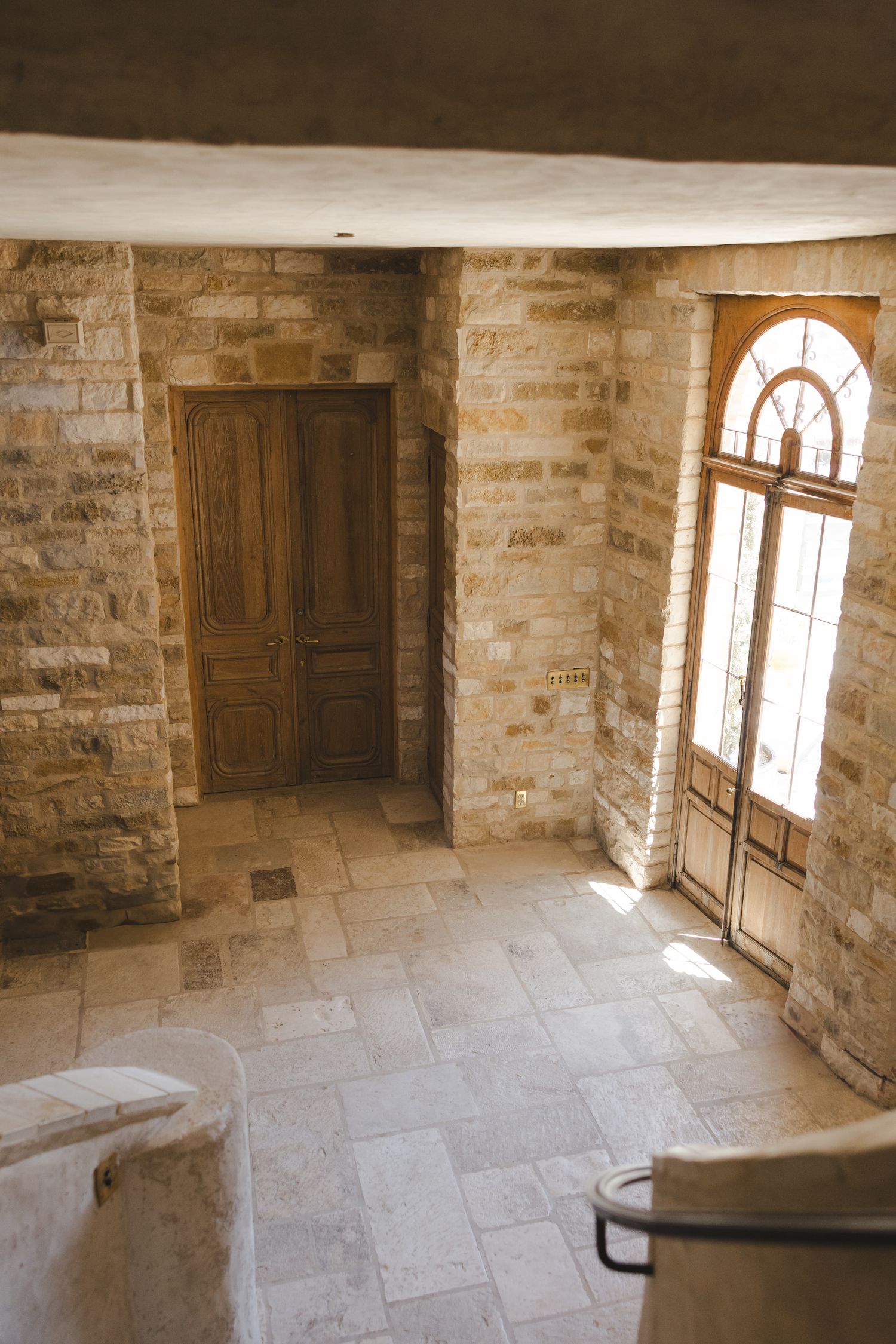 Interior of a rustic stone building with arched windows and wooden doors letting in natural light.