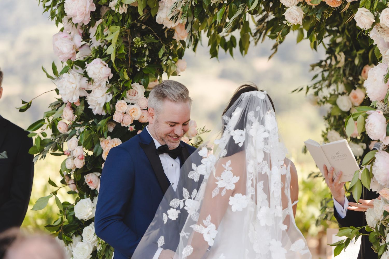 Wedding ceremony under floral arch decorated with pink and white roses in wine country.
