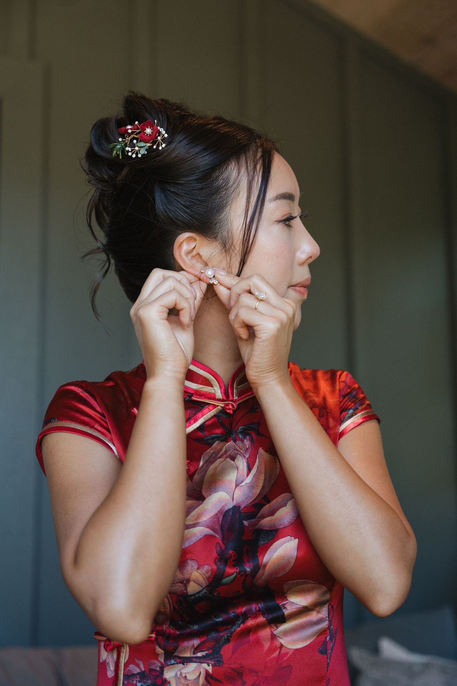 A person wearing a red floral qipao dress puts on earrings while standing in front of a green wall.