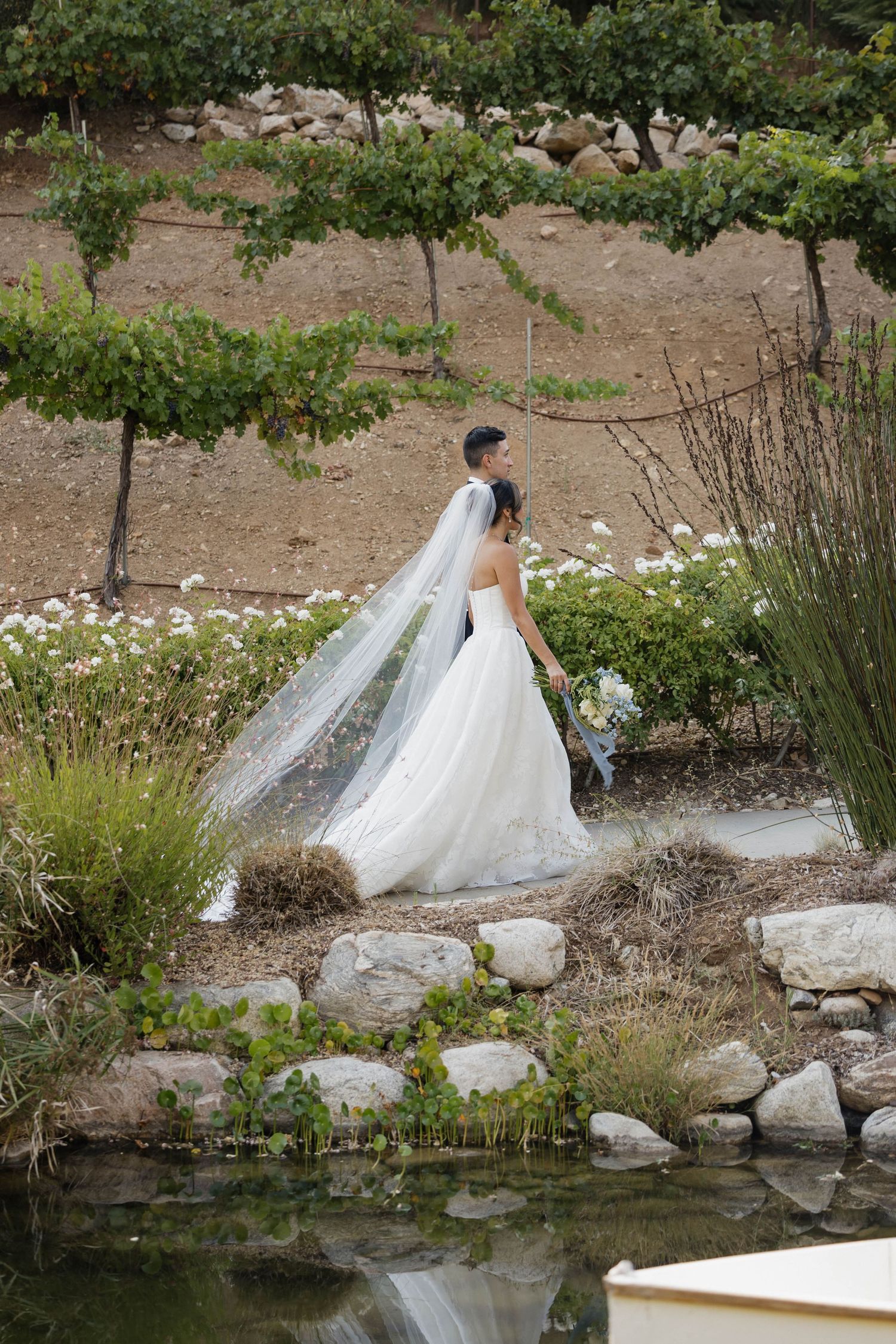 Wedding dress train flows elegantly along garden path with pond and rocks in natural setting.