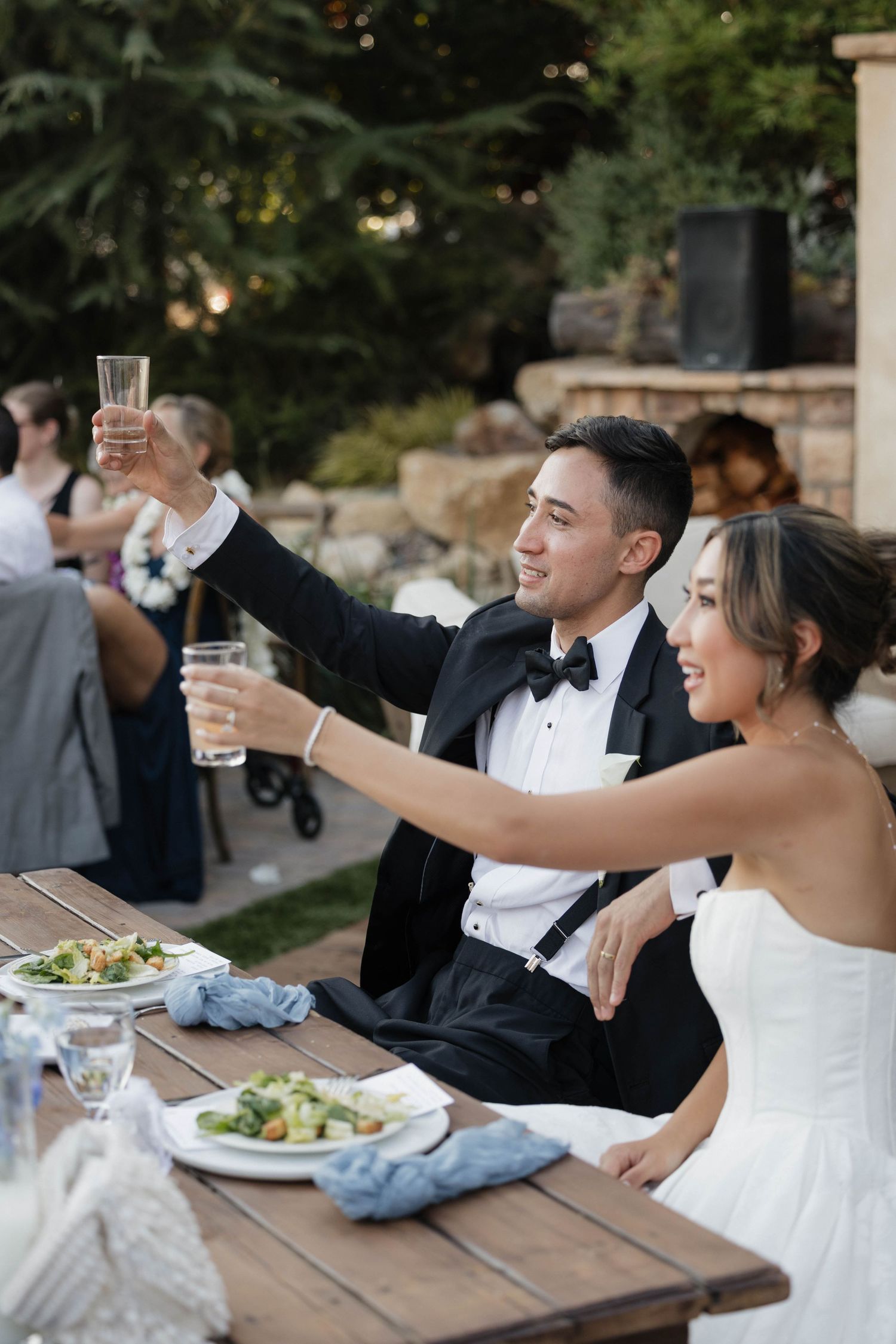 Wedding couple raises champagne glasses in celebration during outdoor reception dinner.