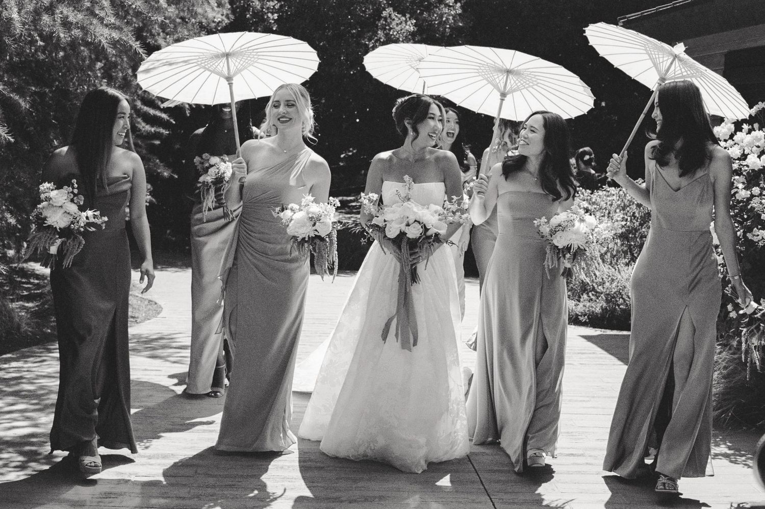 Wedding party participants hold delicate white parasols while standing in a garden during an outdoor ceremony.
