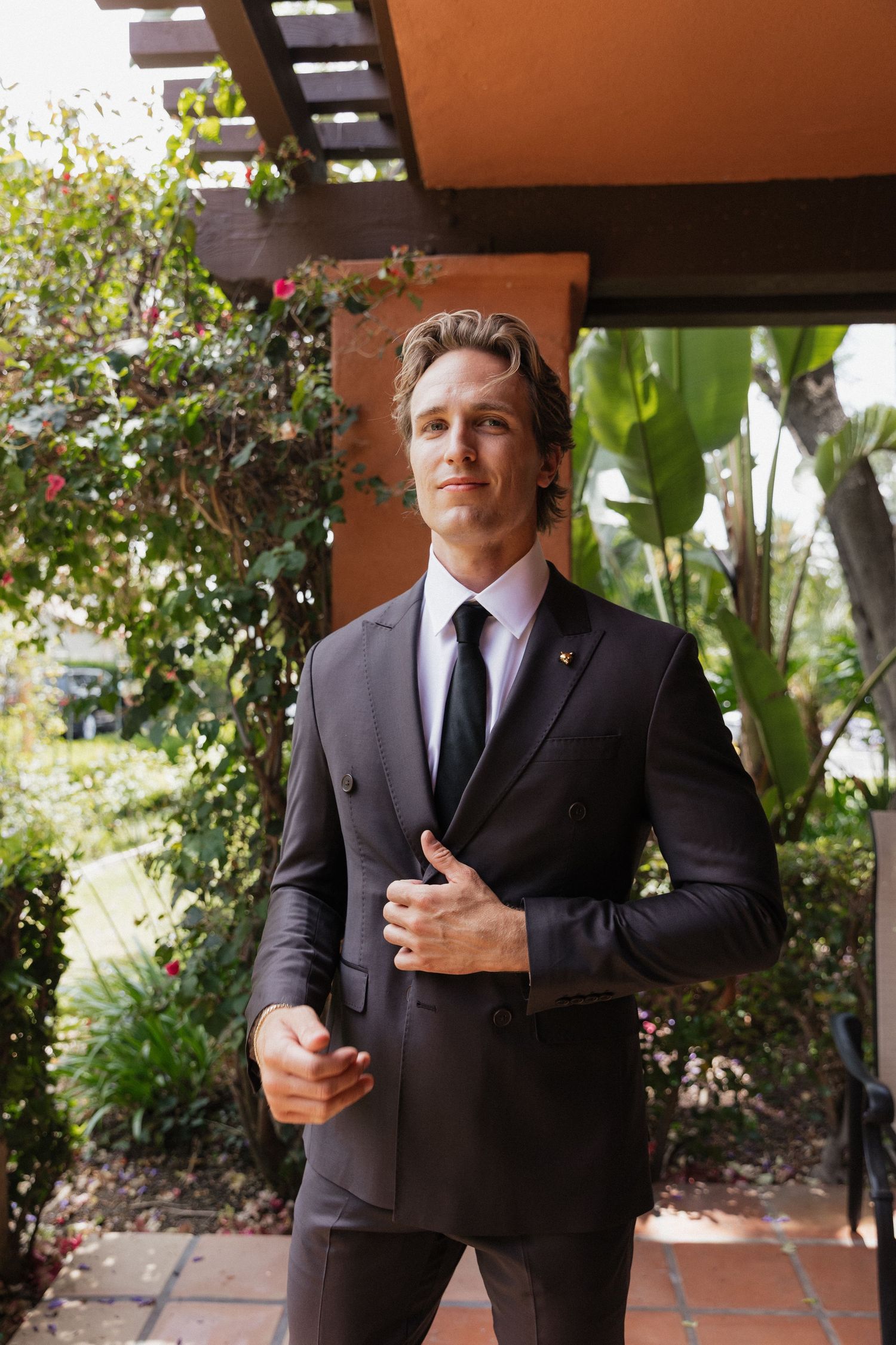 Handsome groom in dark suit and black tie adjusts jacket while standing in garden pergola before wedding.