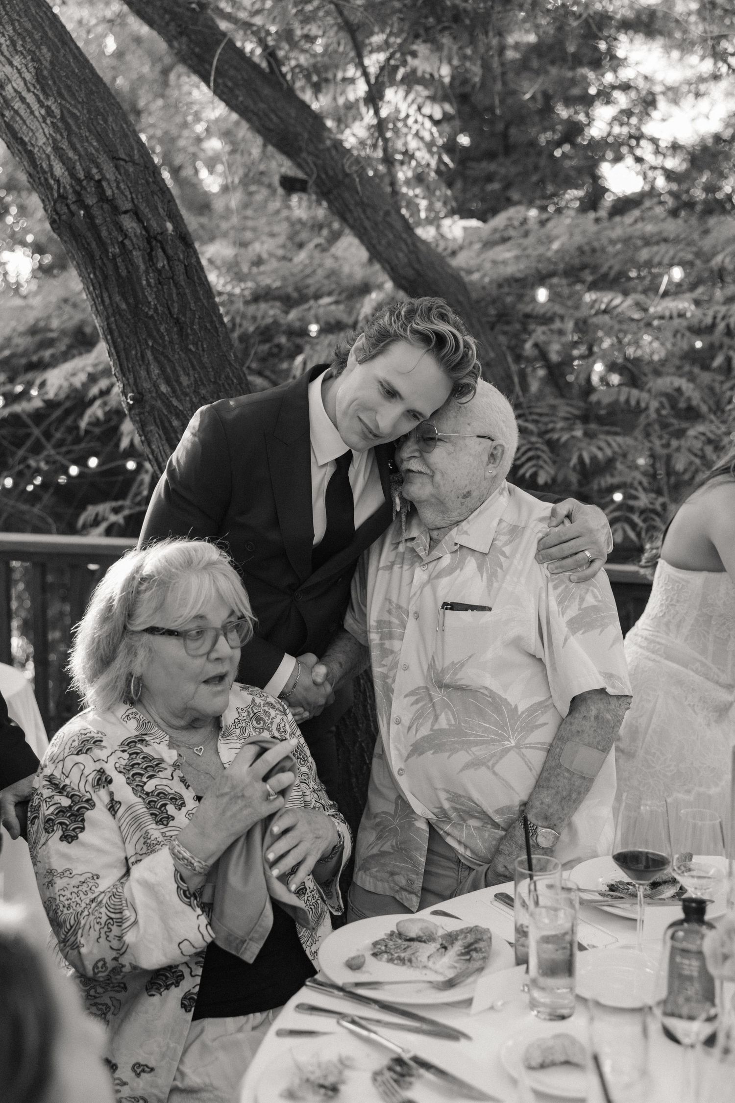 Groom tenderly embraces elderly grandparents at outdoor wedding reception table in black and white.