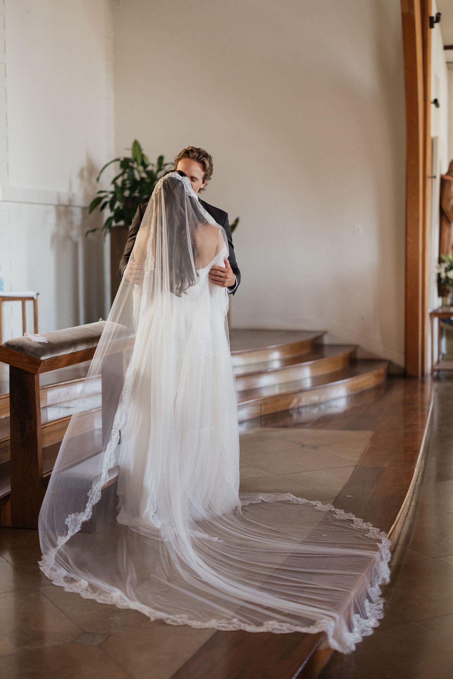 Bride in a flowing gown with a long lace-trimmed veil embraces groom inside a church.