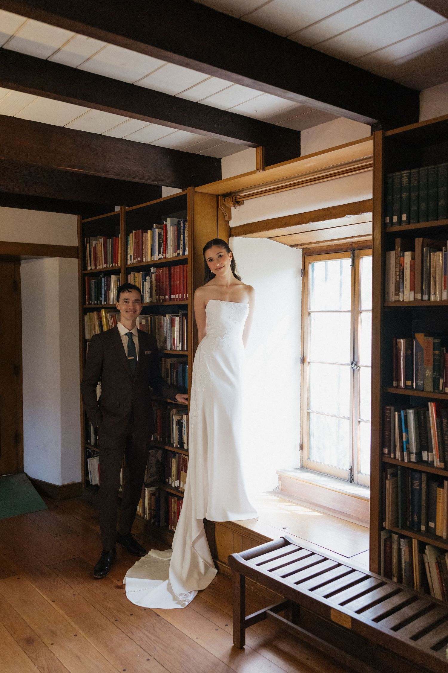 Bride in strapless white gown stands in library window alcove beside suited groom, surrounded by wooden bookshelves.