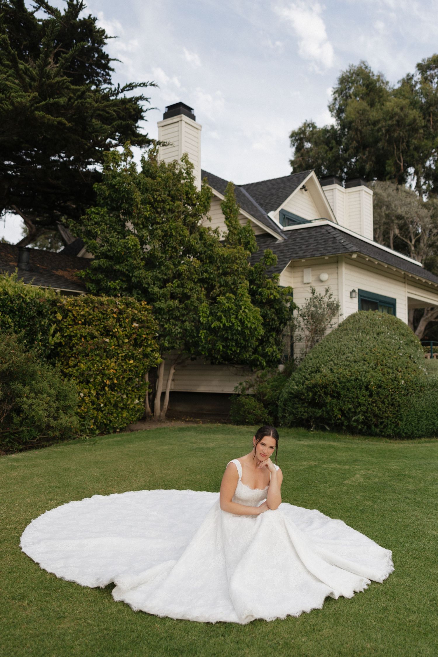 Bride in a flowing white ball gown with long train sits gracefully on green lawn outside elegant estate home.