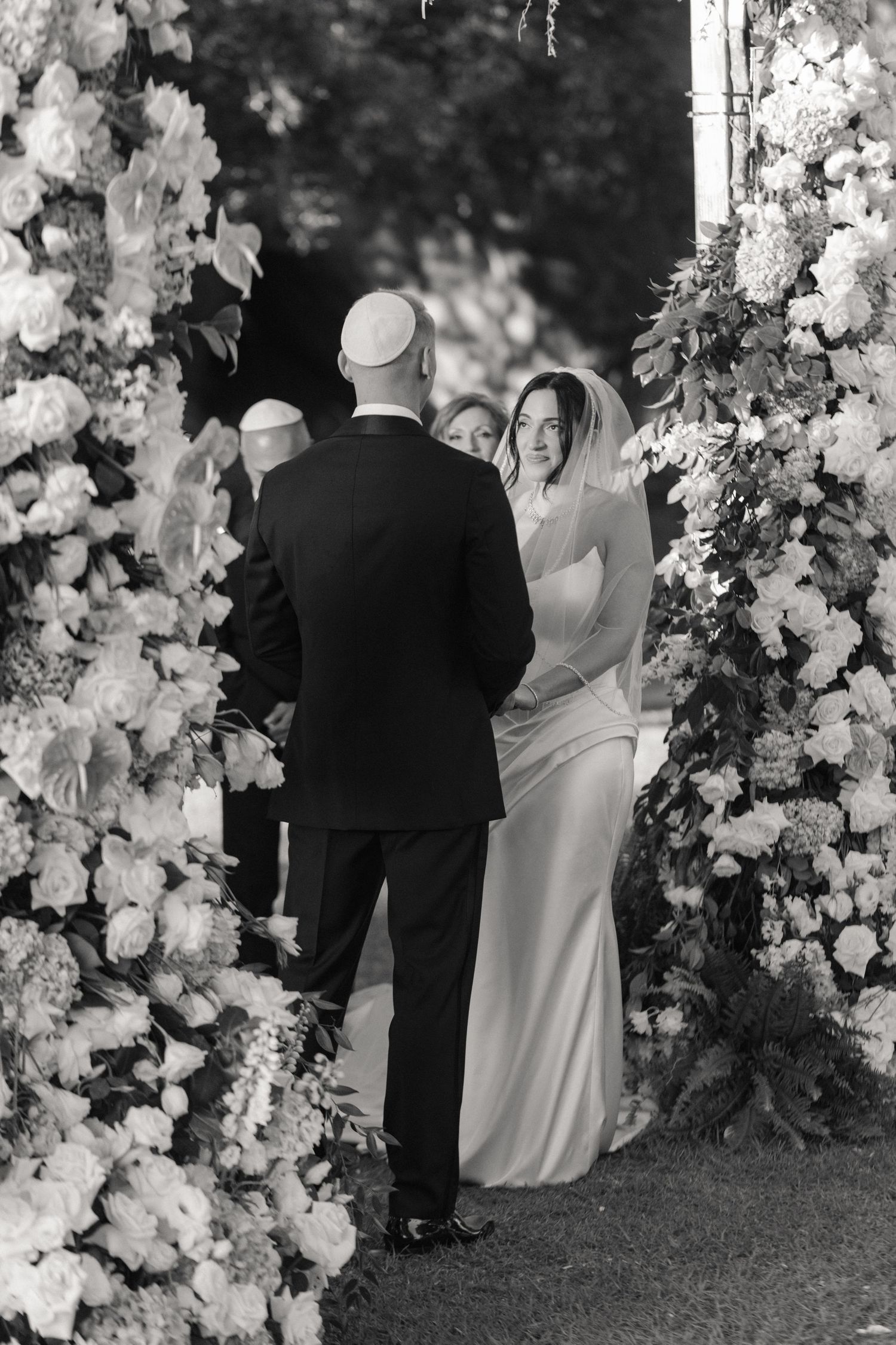 Black and white photo of bride and groom standing at floral wedding altar during outdoor ceremony.