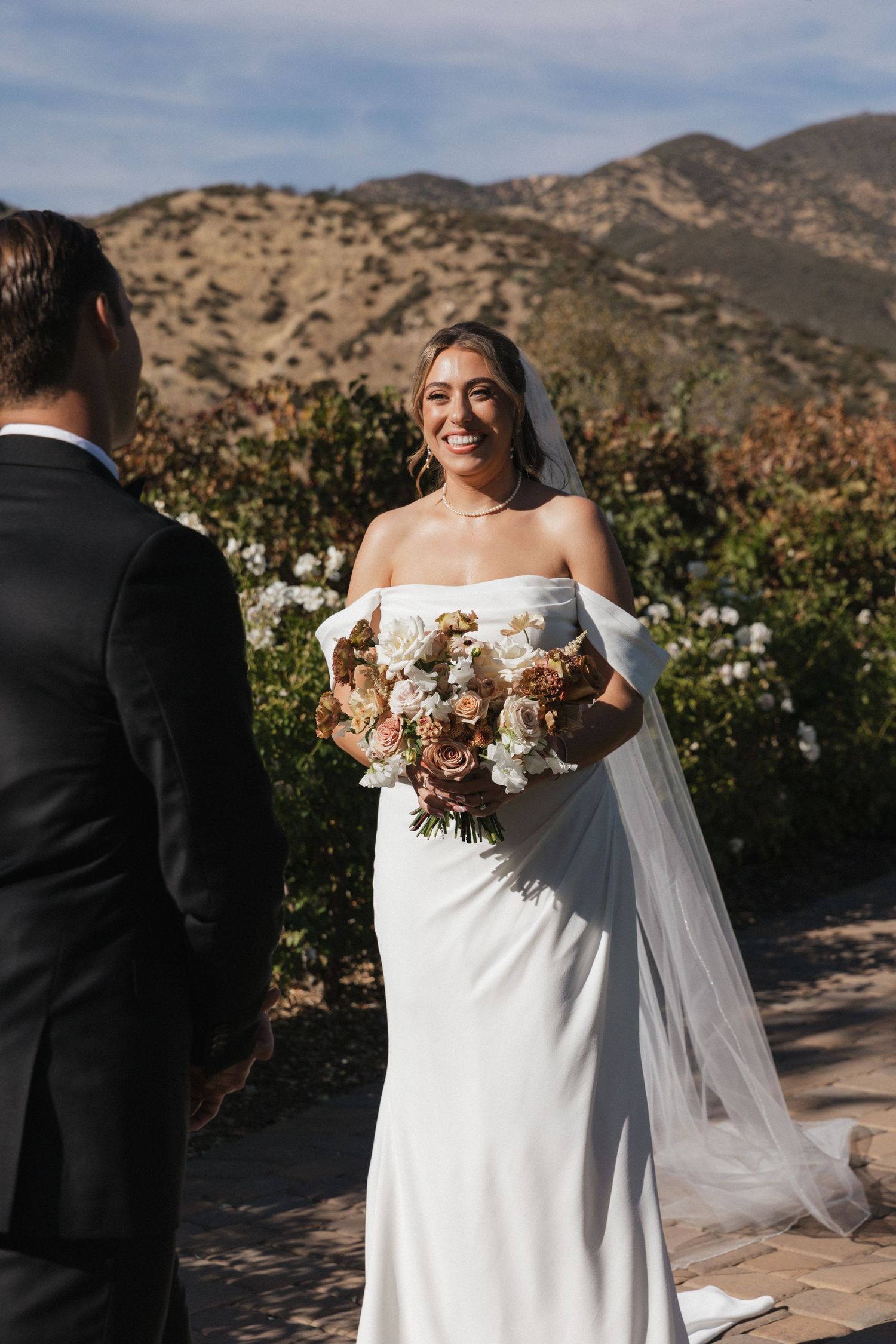 Wedding couple poses against mountainous backdrop with bouquet of dried flowers and white dress.