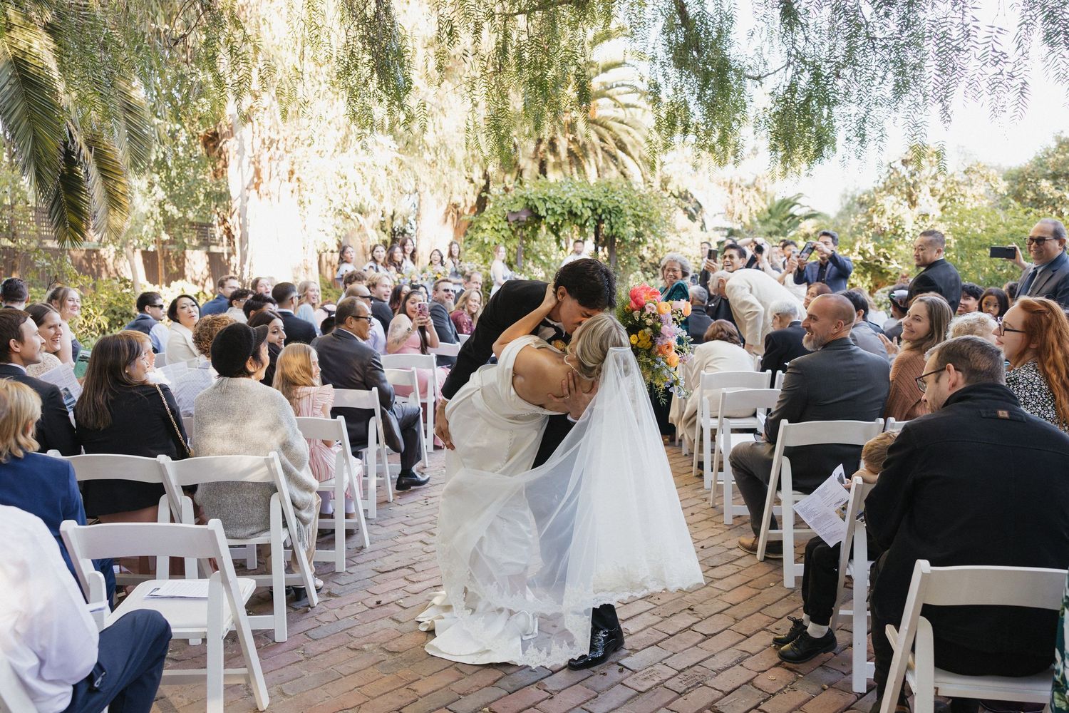 A romantic kiss during a garden wedding ceremony with guests seated on white chairs under hanging willow trees.