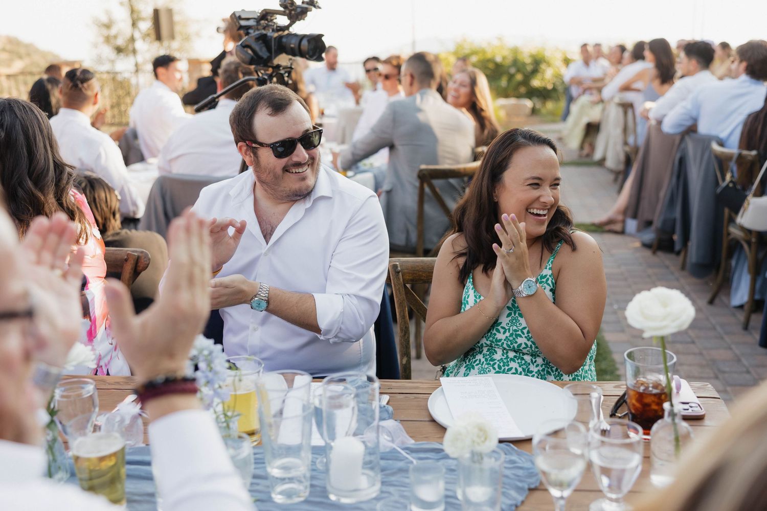 Wedding reception guests enjoying drinks and conversation at outdoor tables with string lights overhead.