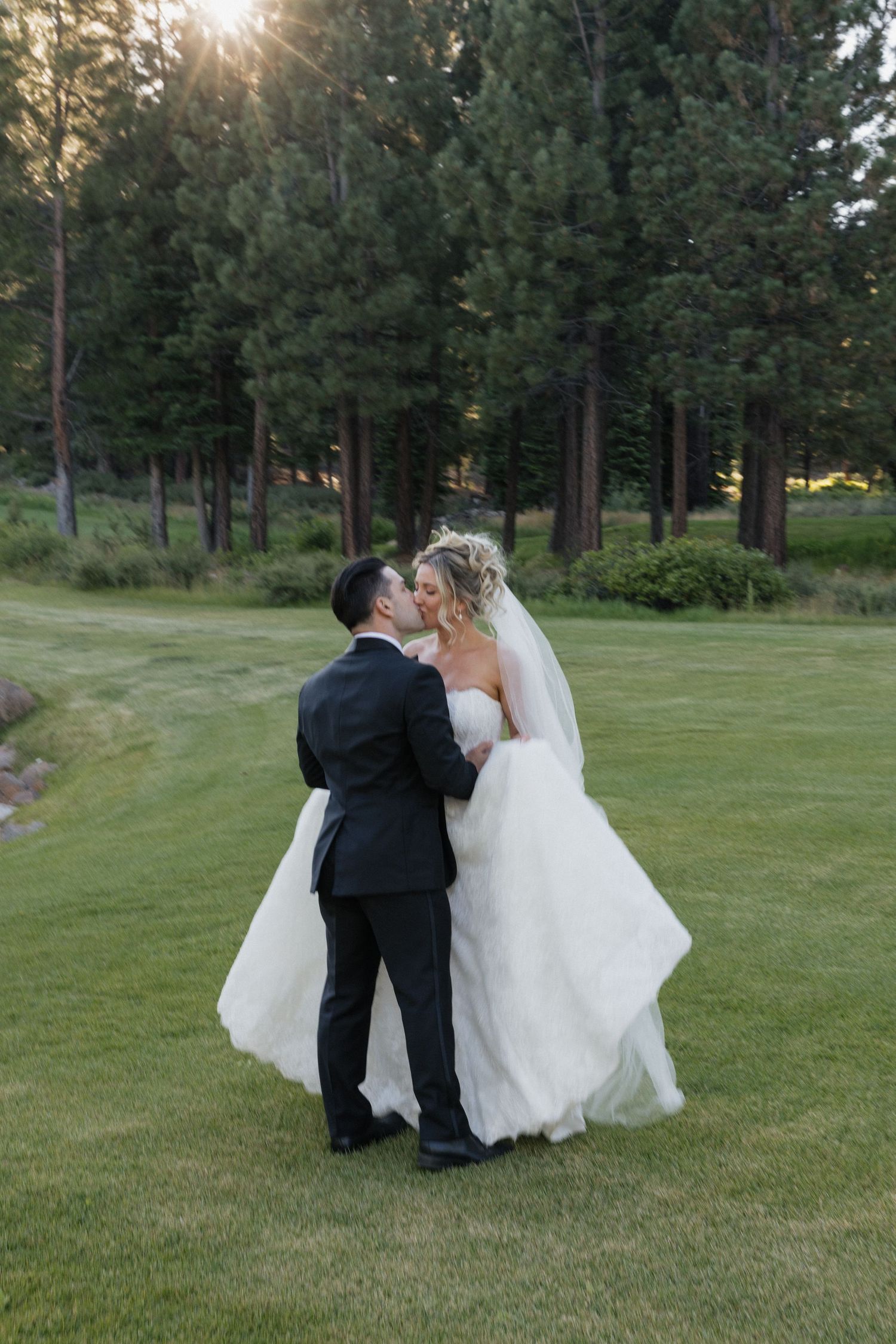 Bride and groom share a romantic kiss on a lush green lawn surrounded by tall pine trees at their outdoor wedding.