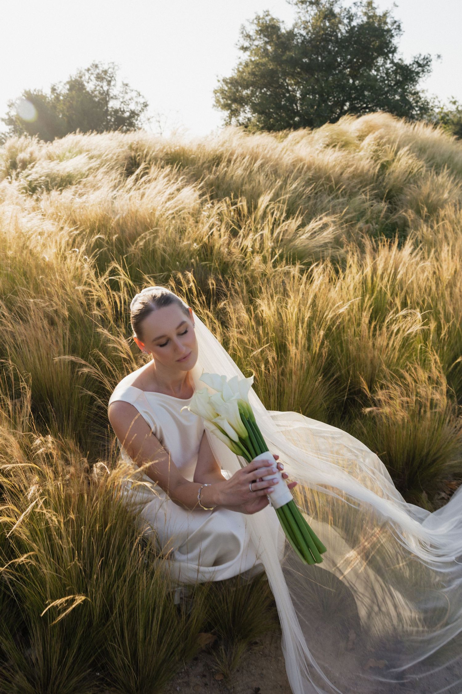 Bride in white gown holding calla lily bouquet sitting in golden grass field with veil flowing in breeze.