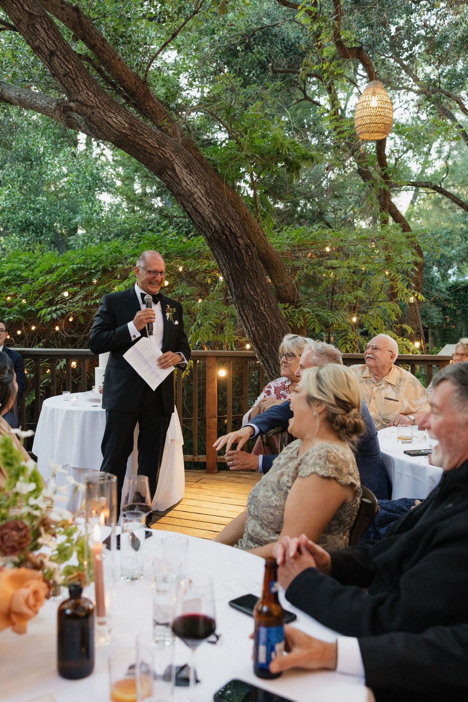 Guest delivers heartfelt speech at outdoor wedding reception under string lights and lush trees.