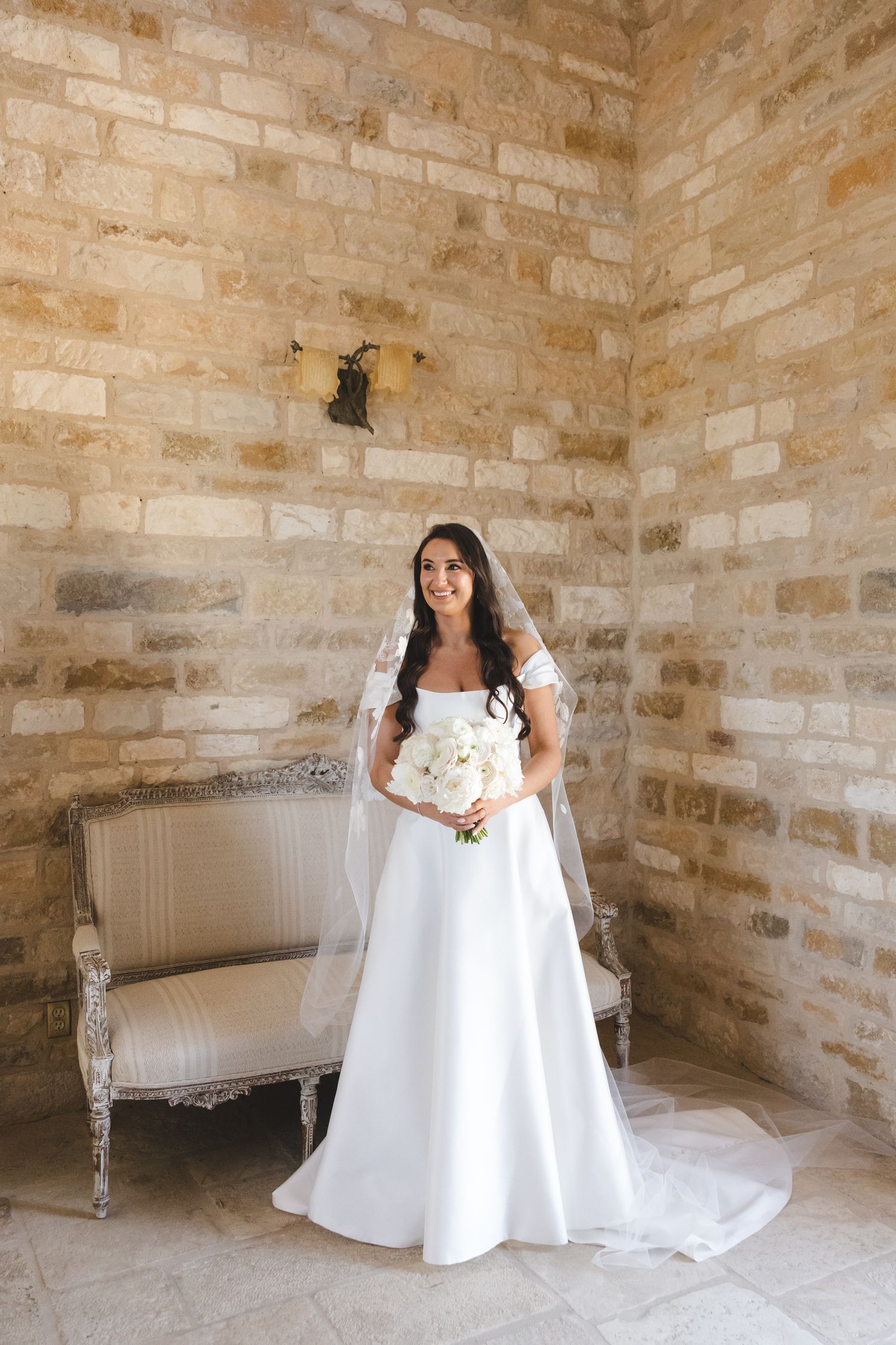 Elegant white wedding dress flows across a vintage chaise lounge in a rustic stone room.