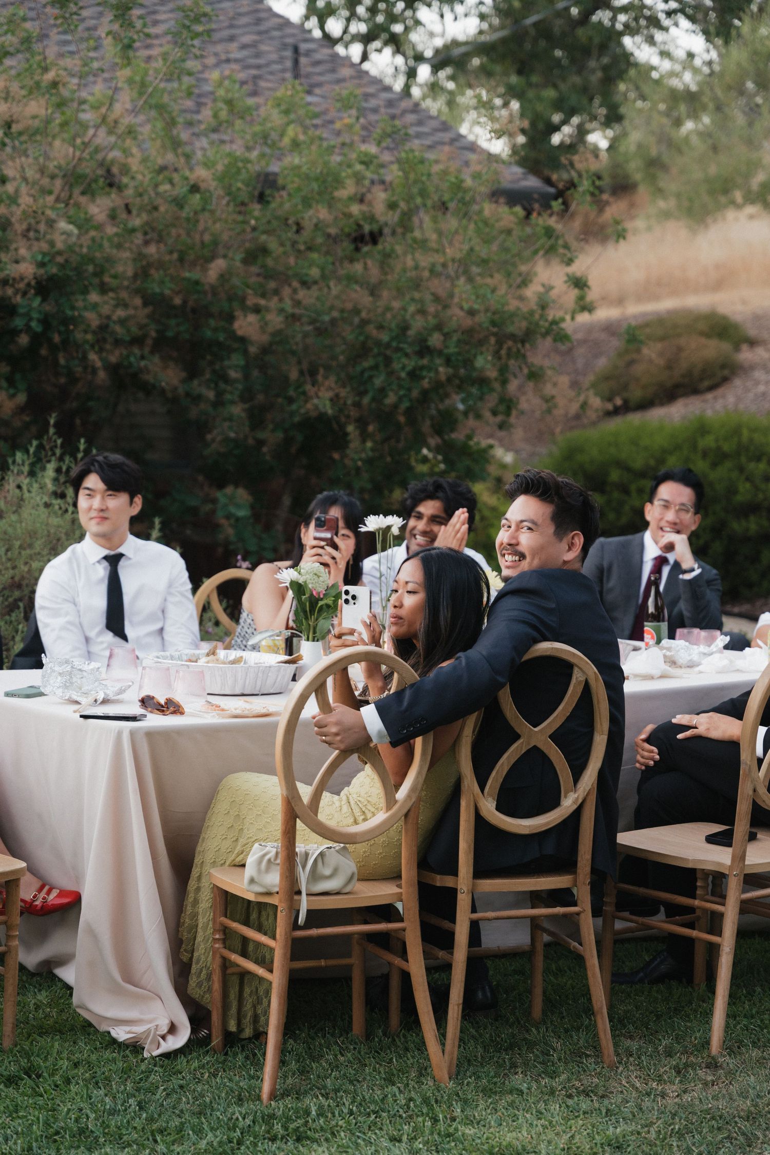 Wedding guests enjoy a casual outdoor dinner at long tables with wooden chairs on a grassy lawn.