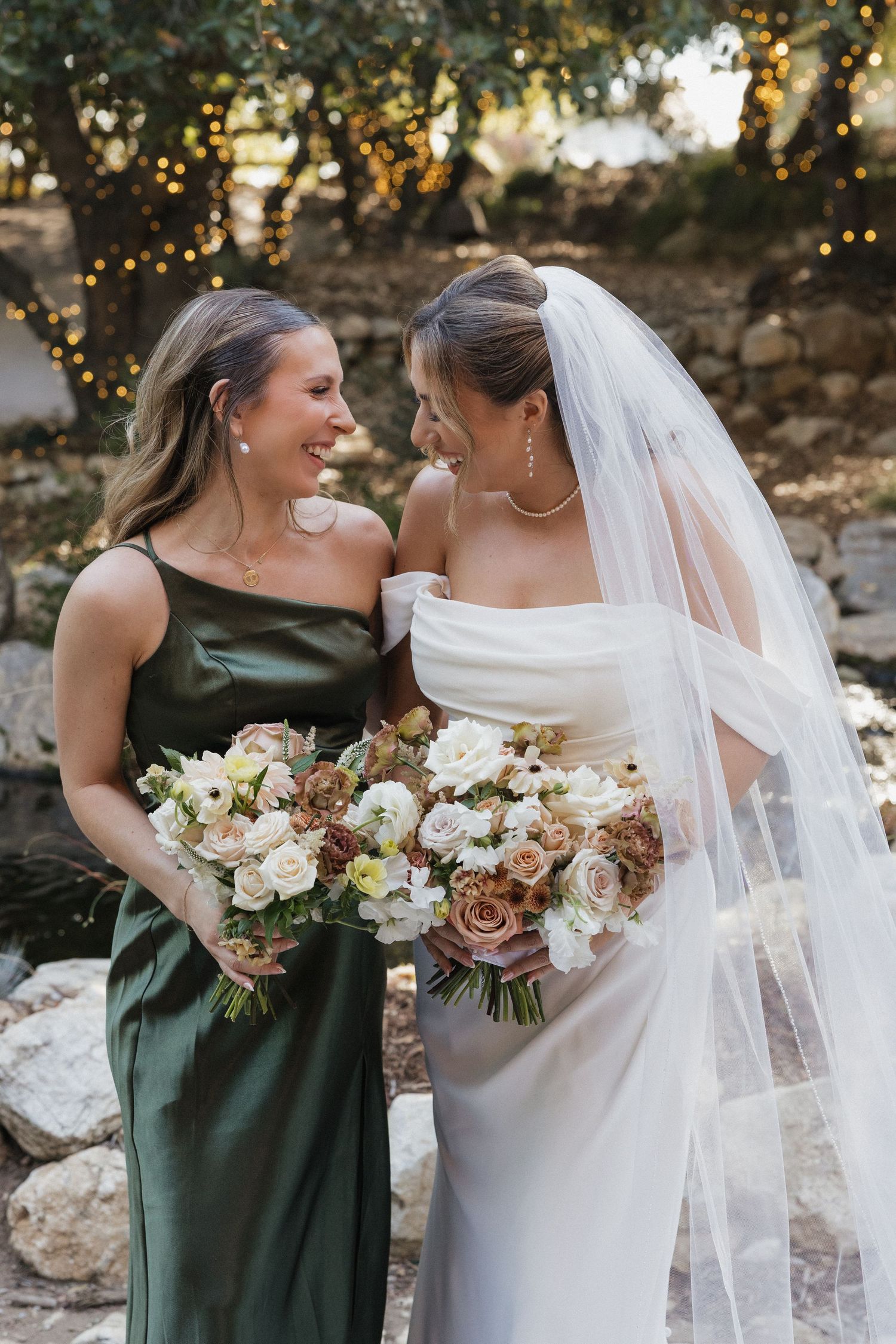 Wedding party poses with bouquets featuring white and green florals against stone wall backdrop.