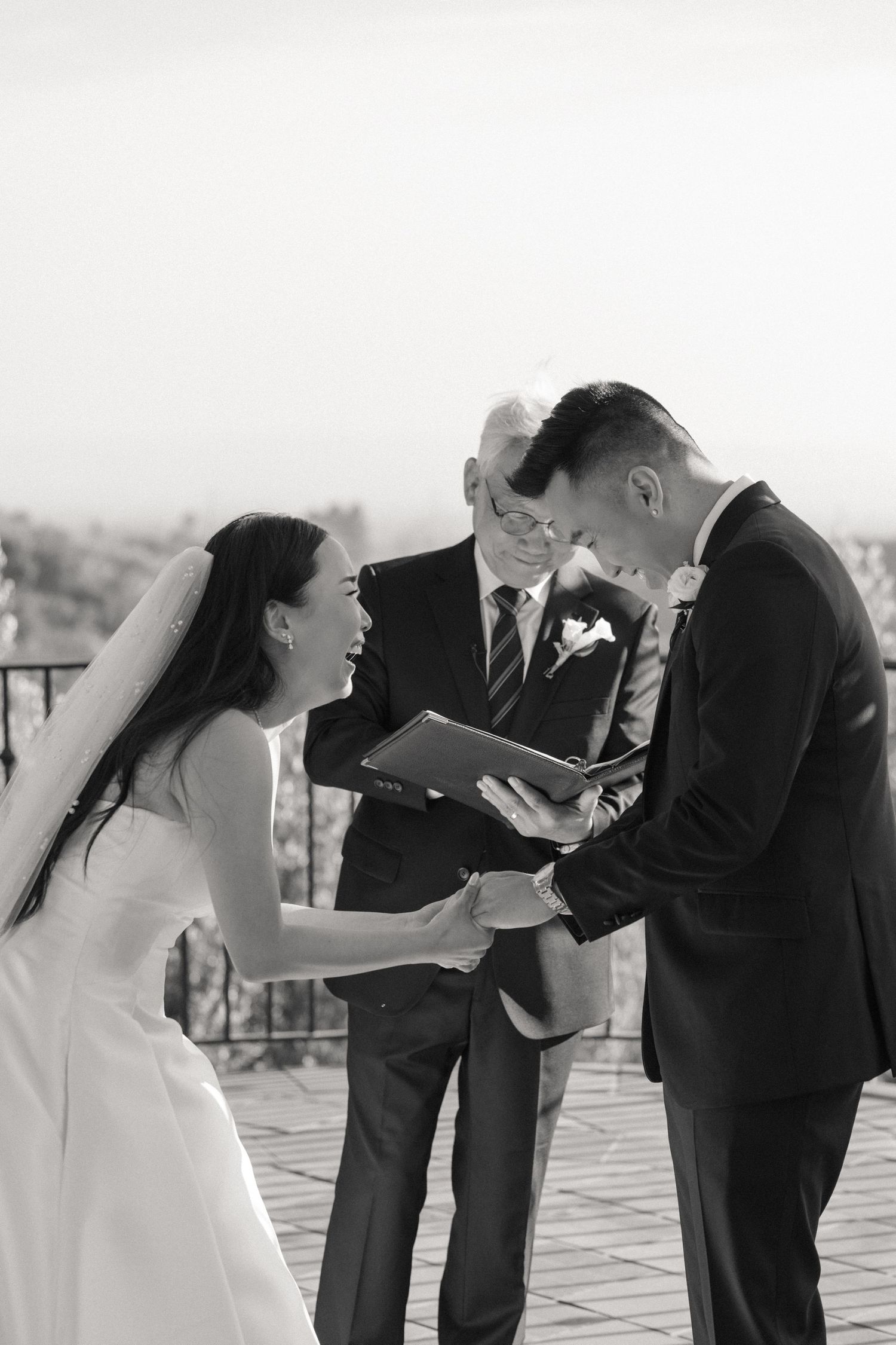 Black and white photo of a bride and groom exchanging vows with an officiant at an outdoor wedding ceremony.