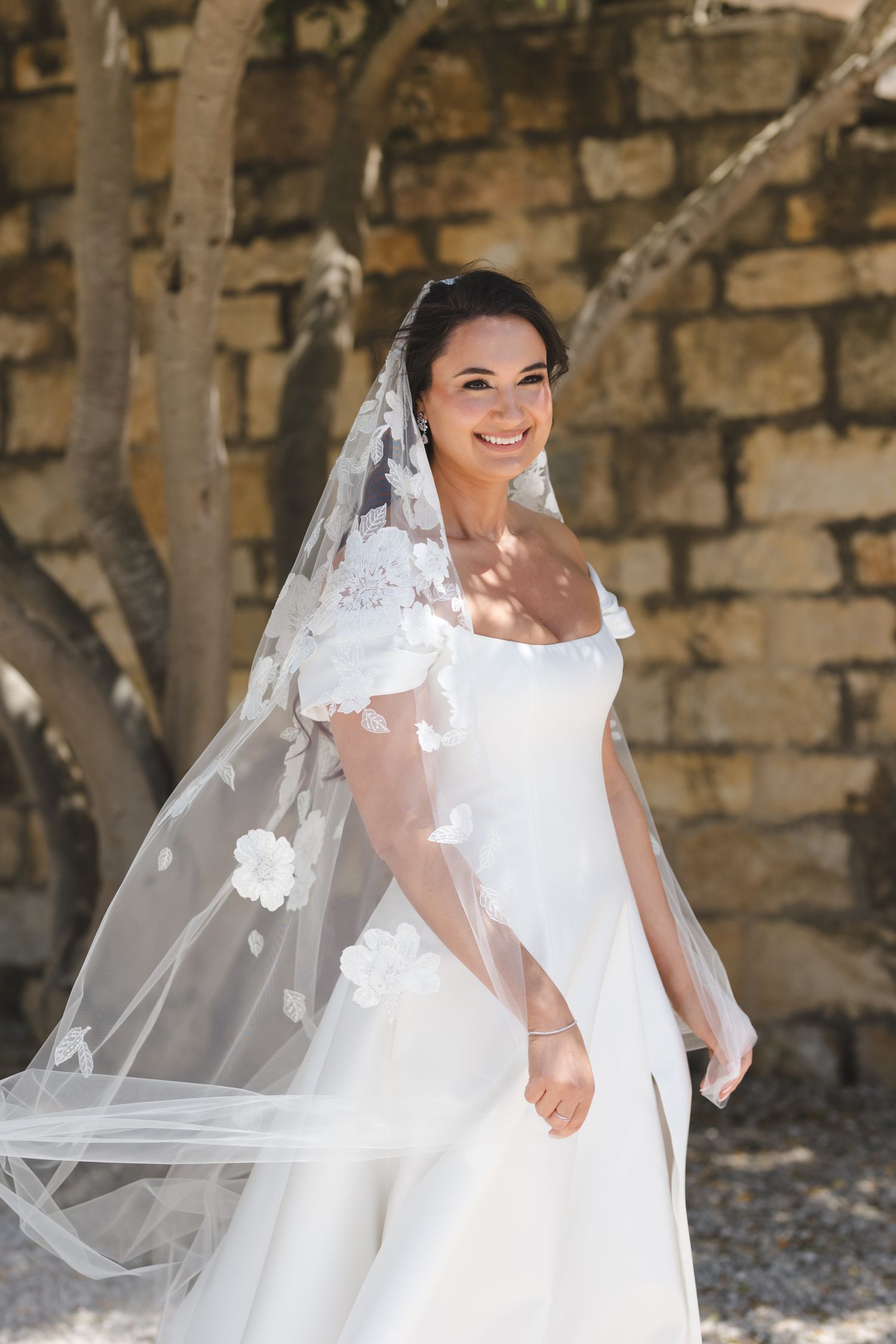 Bride in elegant white gown and veil poses against stone wall in dappled sunlight.